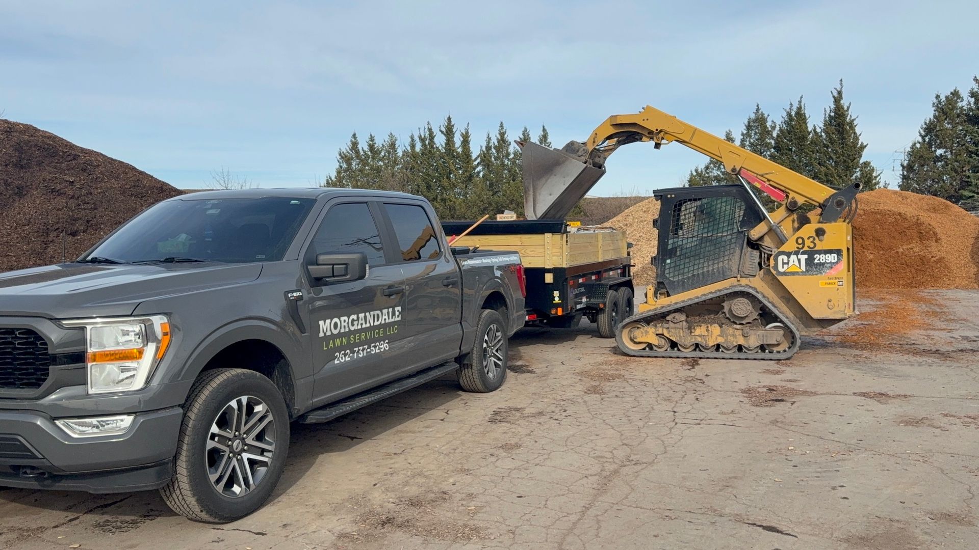 Black pickup truck beside a yellow wood chipper or excavator at a gravel yard under trees