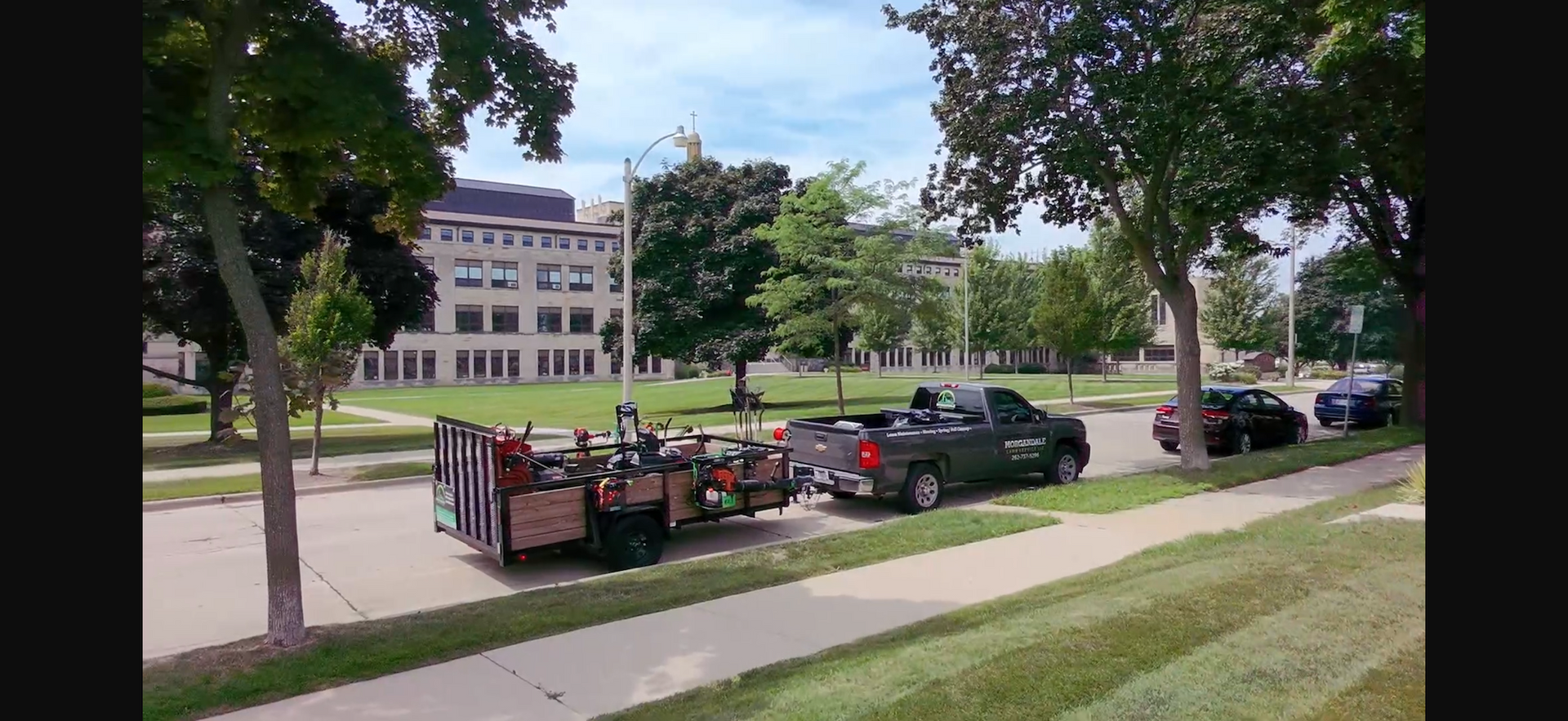 Parked vehicles and a red dumpster beside a sidewalk in front of a grassy campus building with trees.