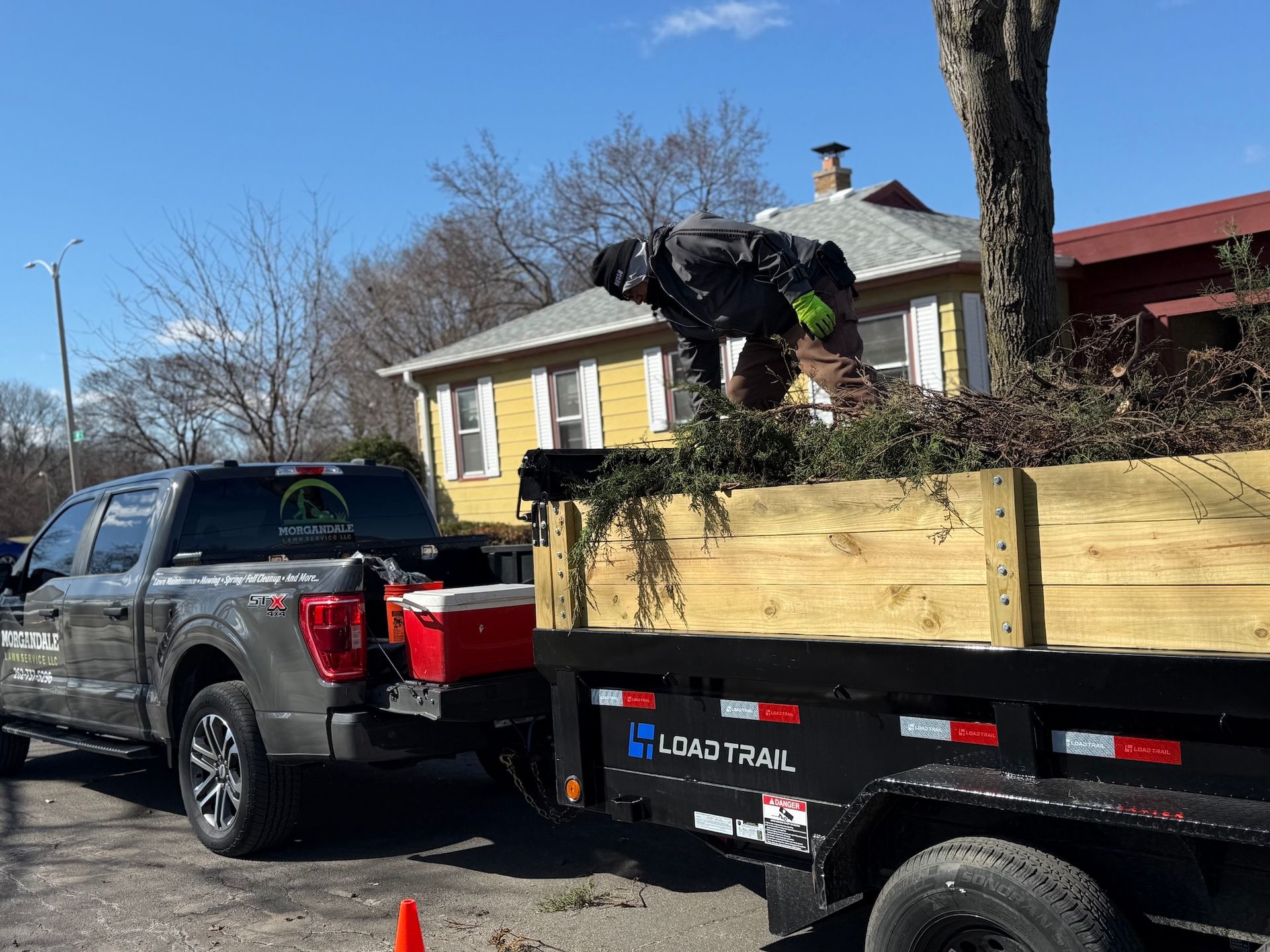 Worker loading branches into a trailer hitched to a black pickup truck on a residential street