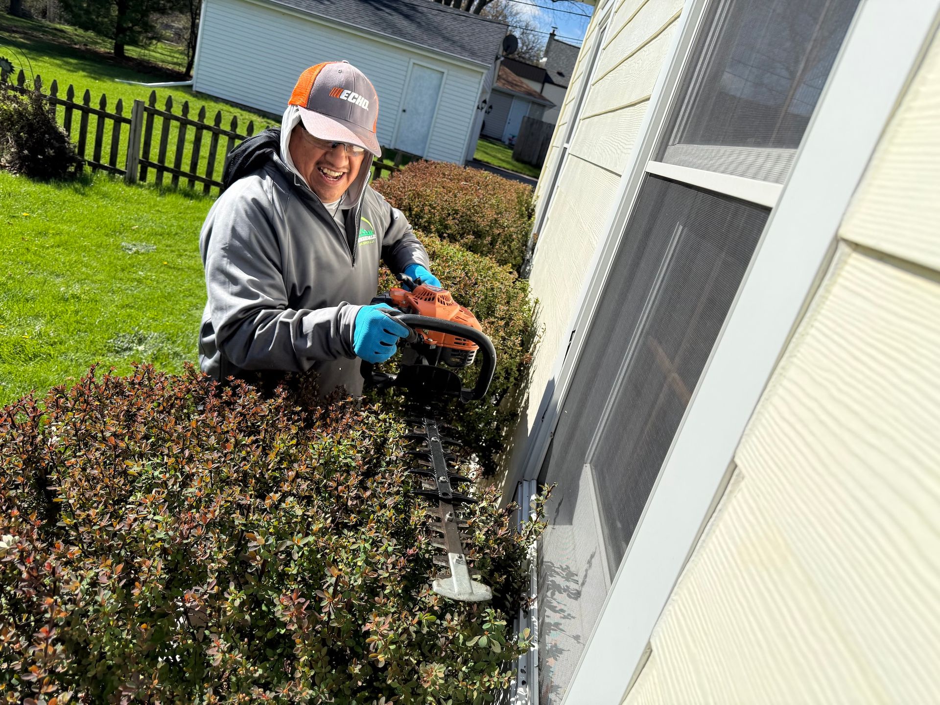 Worker trimming bushes beside a house, wearing a helmet and blue gloves.