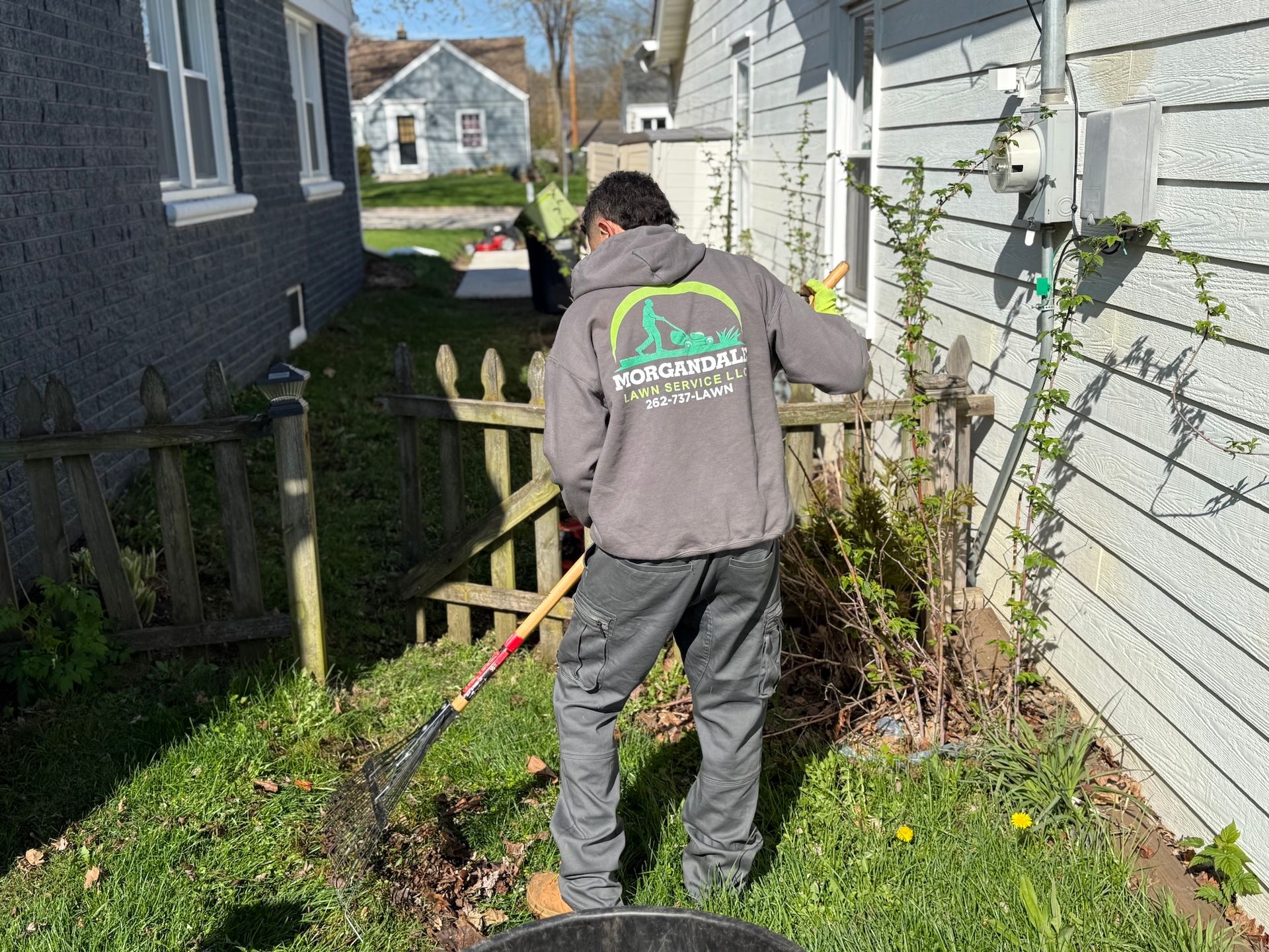 Person using a string trimmer in a narrow grassy side yard beside two houses.