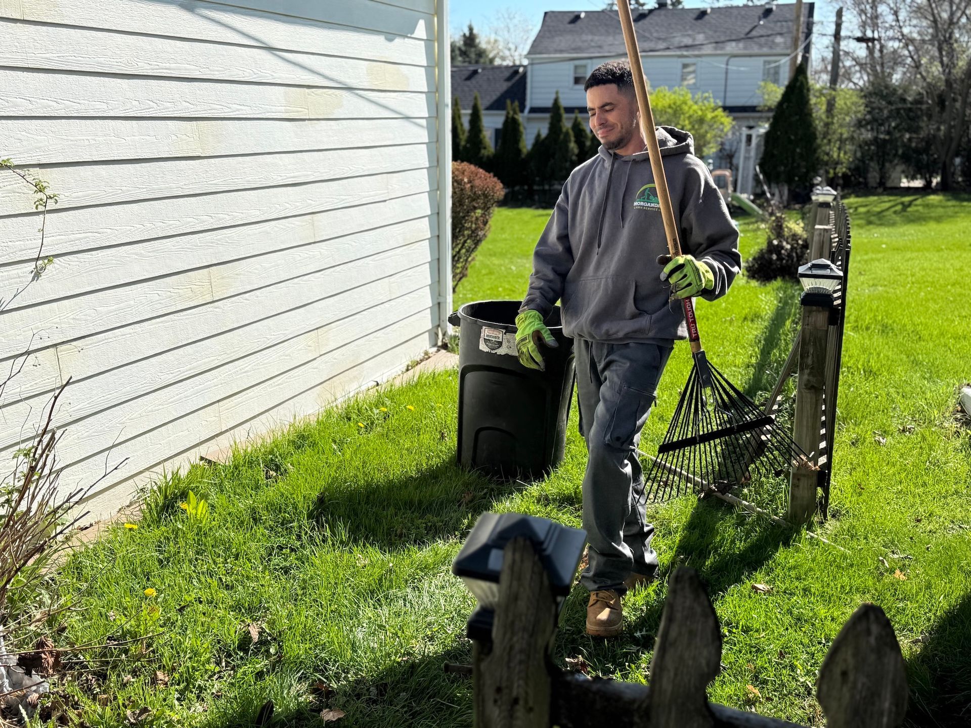 Man standing on a grassy side yard beside a white house, holding a shovel and bucket near a bicycle.