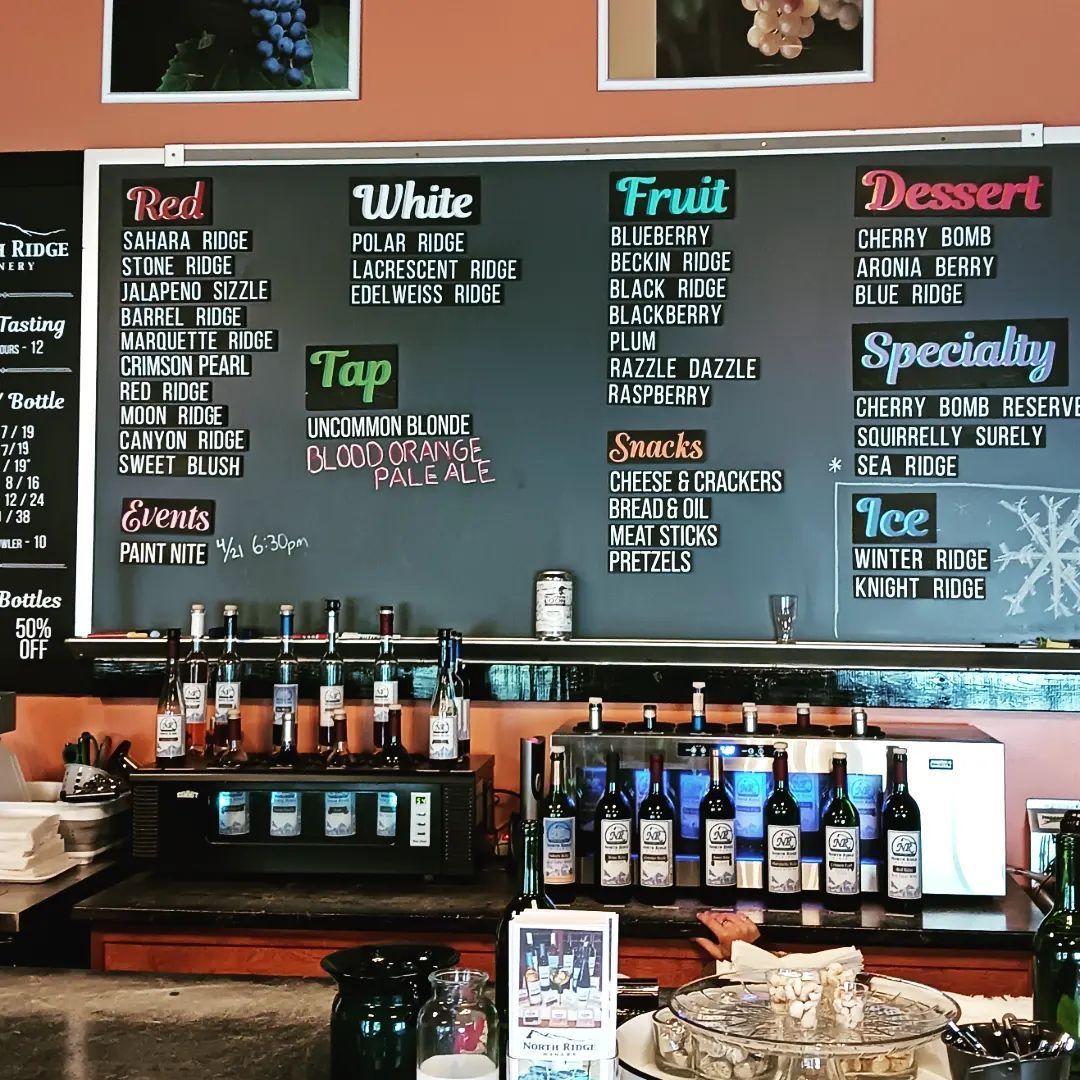 A menu board in a winery lists varieties of red, white, fruit, and dessert wines, plus specialty drinks, snacks, and events.