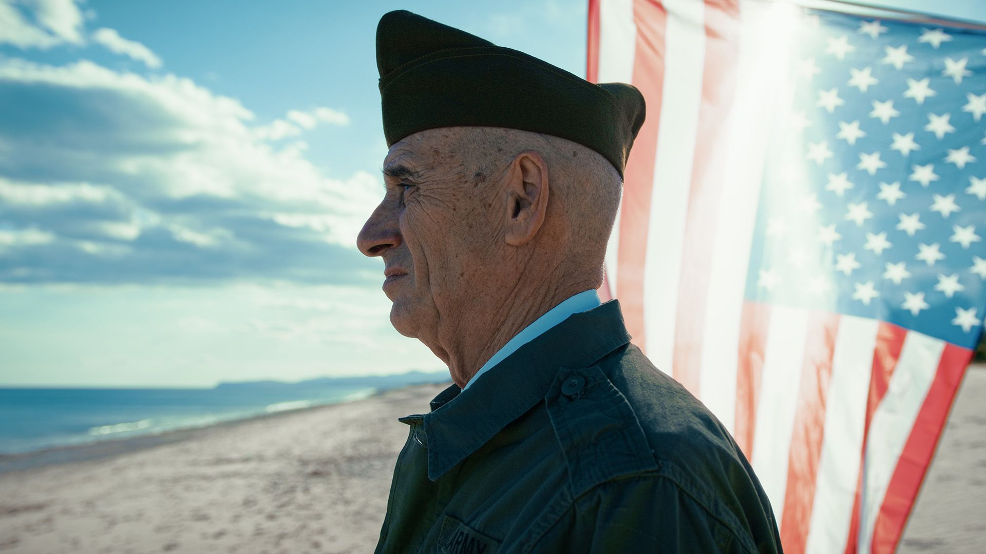 Veteran in uniform stands on beach with American flag.