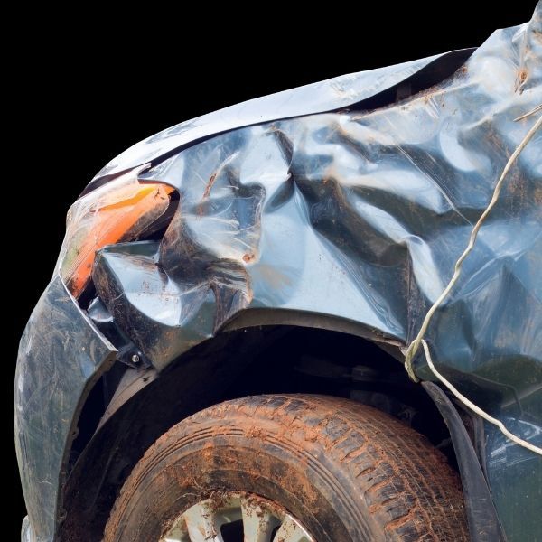 A dark-colored car with severe damage to the front fender and wheel well, covered in mud against a black background.