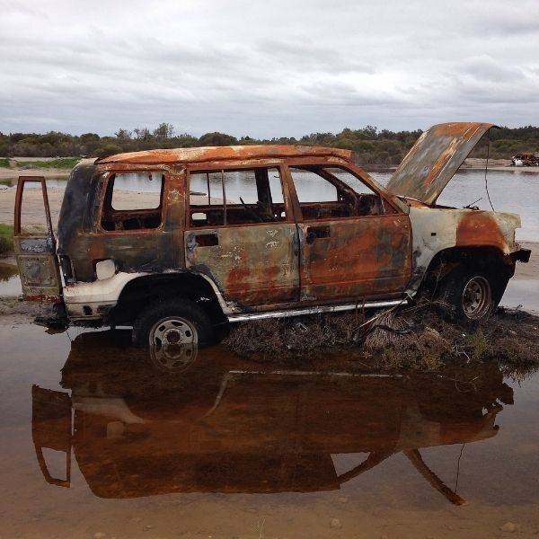 A rusted, burned-out SUV sits partially submerged in a shallow, murky pool under an overcast sky.