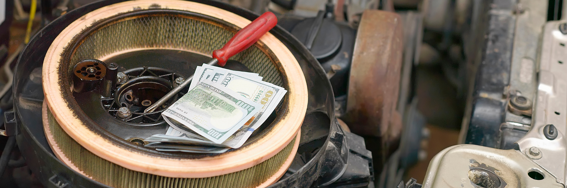 A screwdriver and a stack of US dollar bills sit on top of a circular engine air filter inside an open car engine bay.