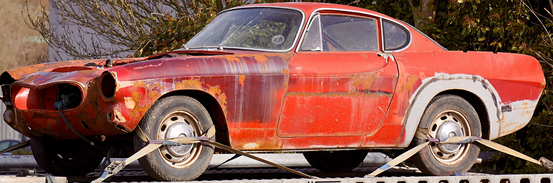 A rusted red vintage sports car secured with straps on a flatbed trailer.