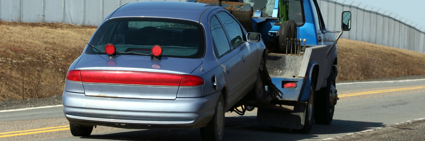 A blue sedan is being towed by a blue tow truck along a highway next to a field.