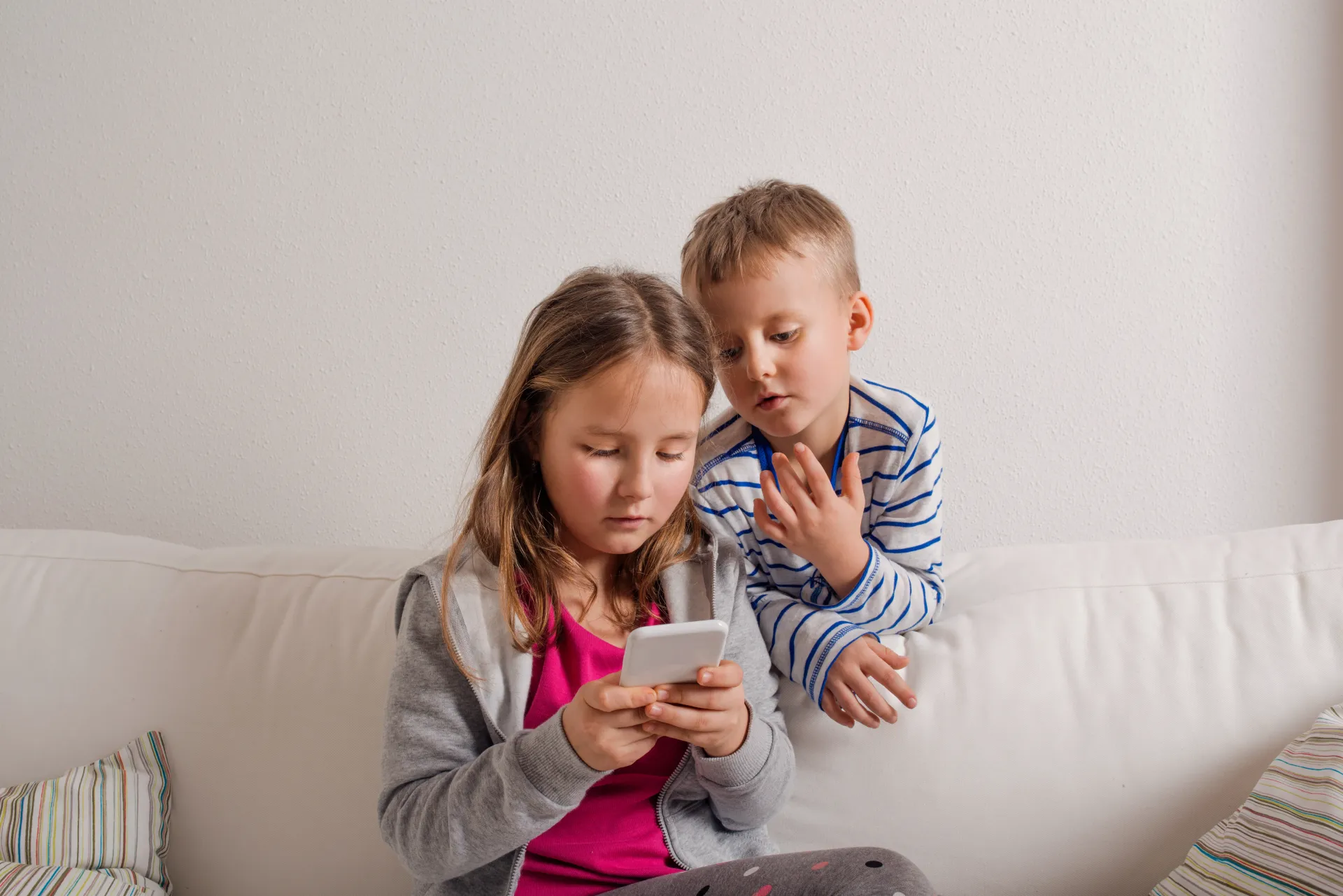 Two children on a white couch: girl looking at phone, boy leaning over her shoulder.