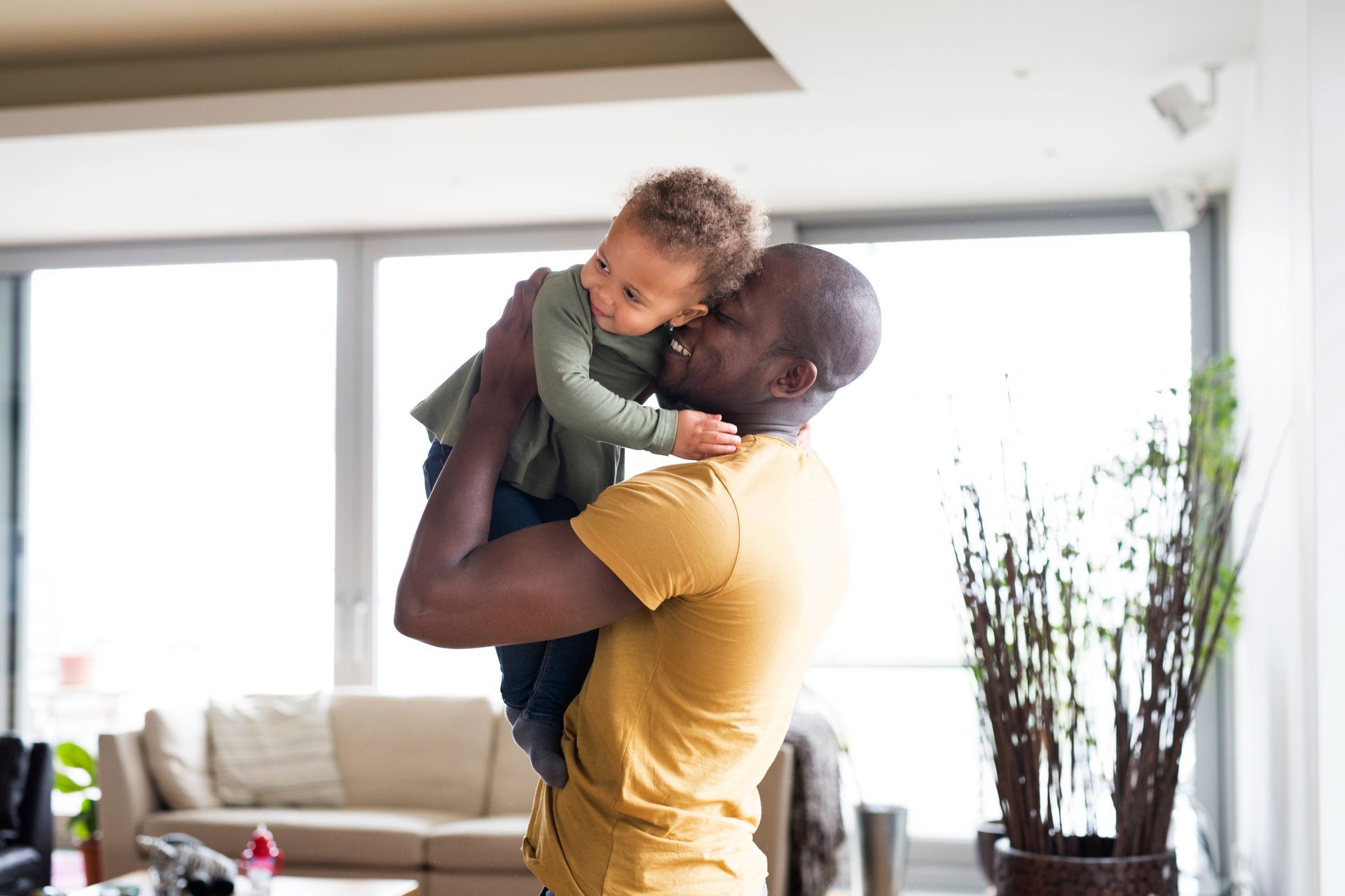 Man holding child indoors, both smiling. Bright room, large windows.