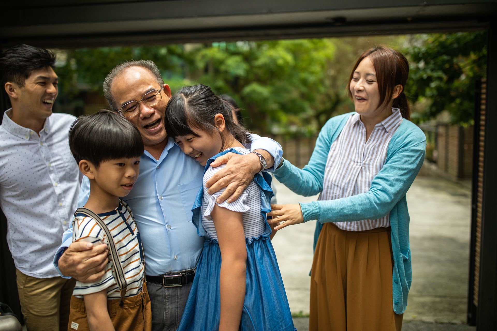 Family embraces in front of a home; grandfather hugs grandchildren, parents smile.