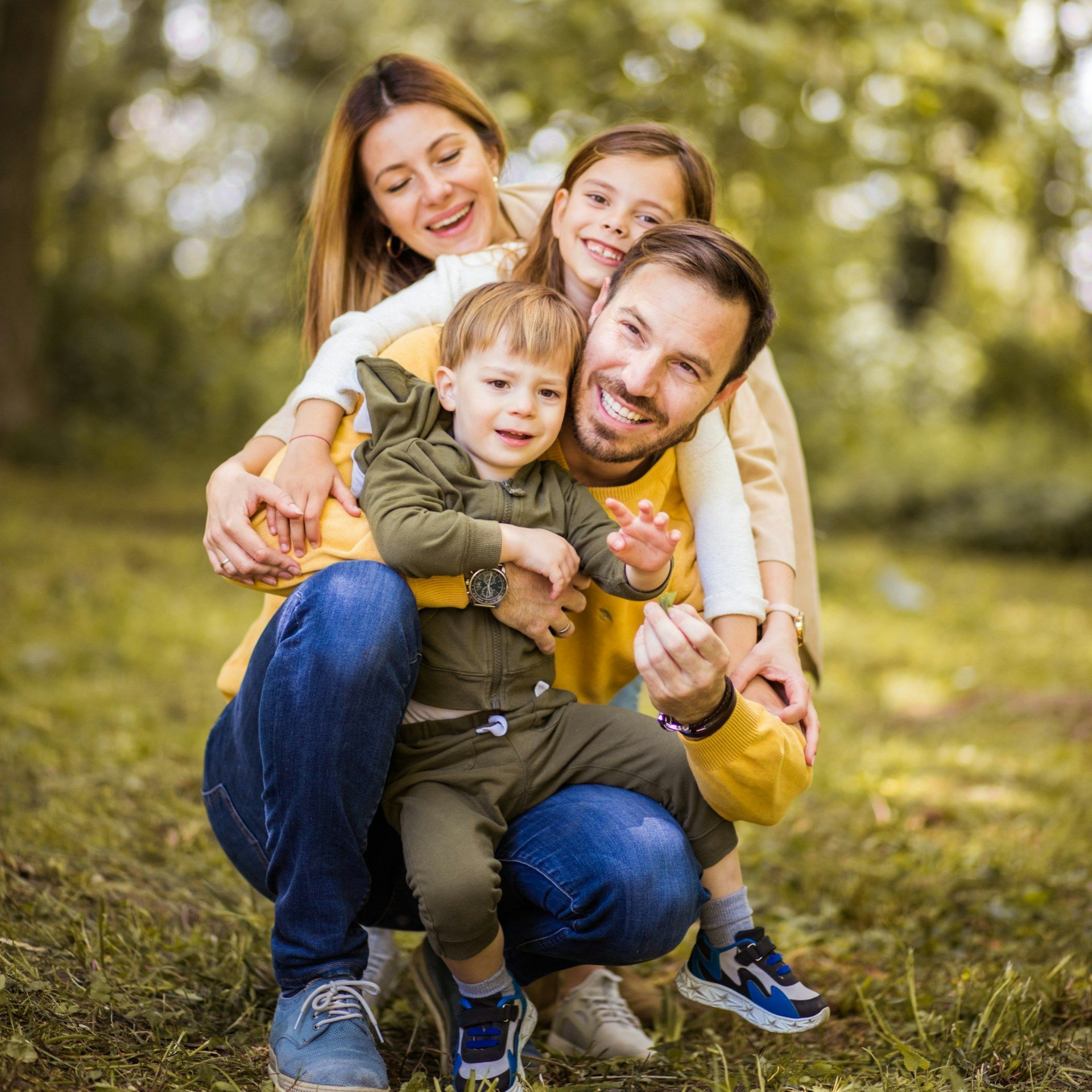 Smiling family of four hugging outdoors in a sunlit park with blurred trees behind them