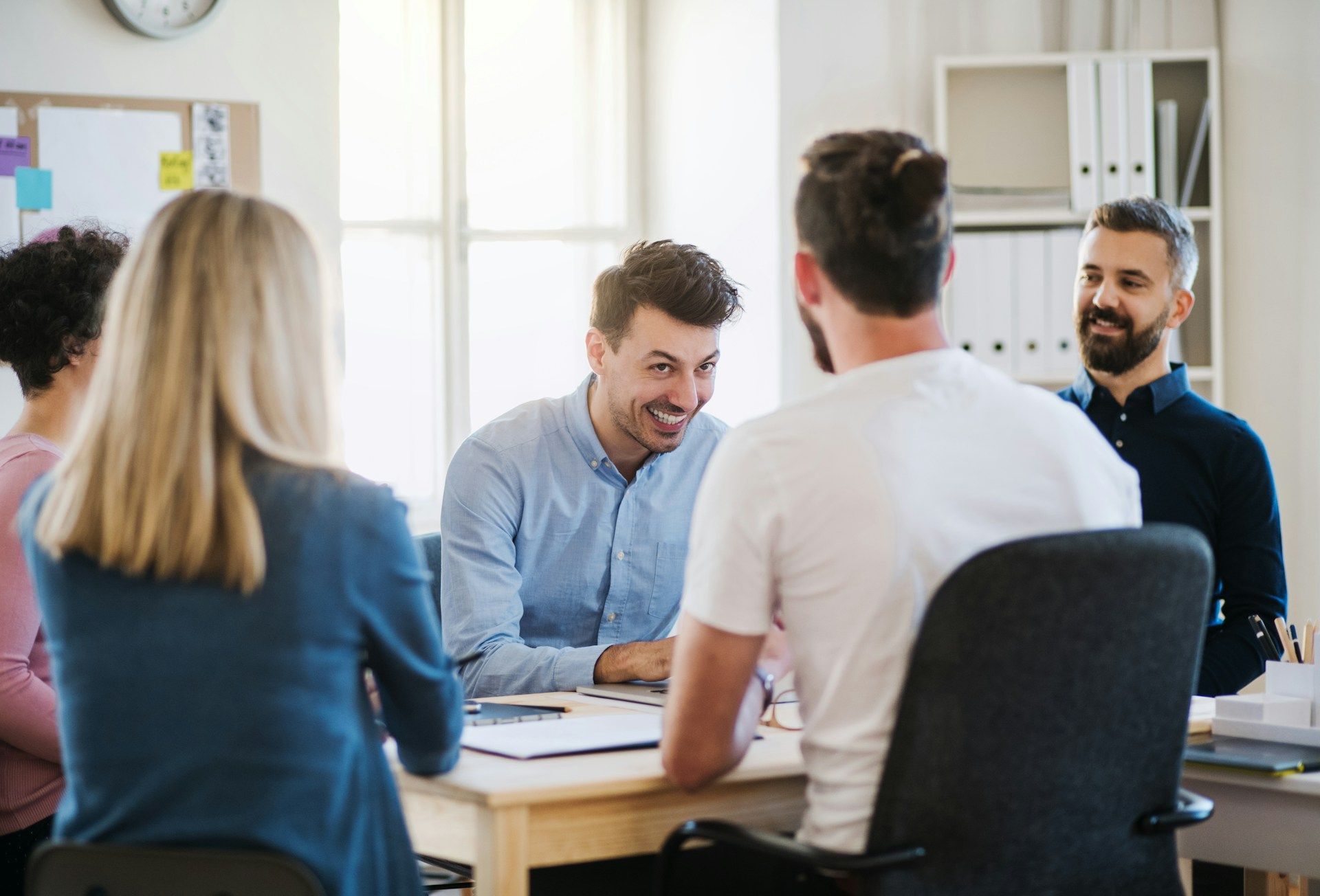 People in a meeting around a table in a bright office, talking and smiling.