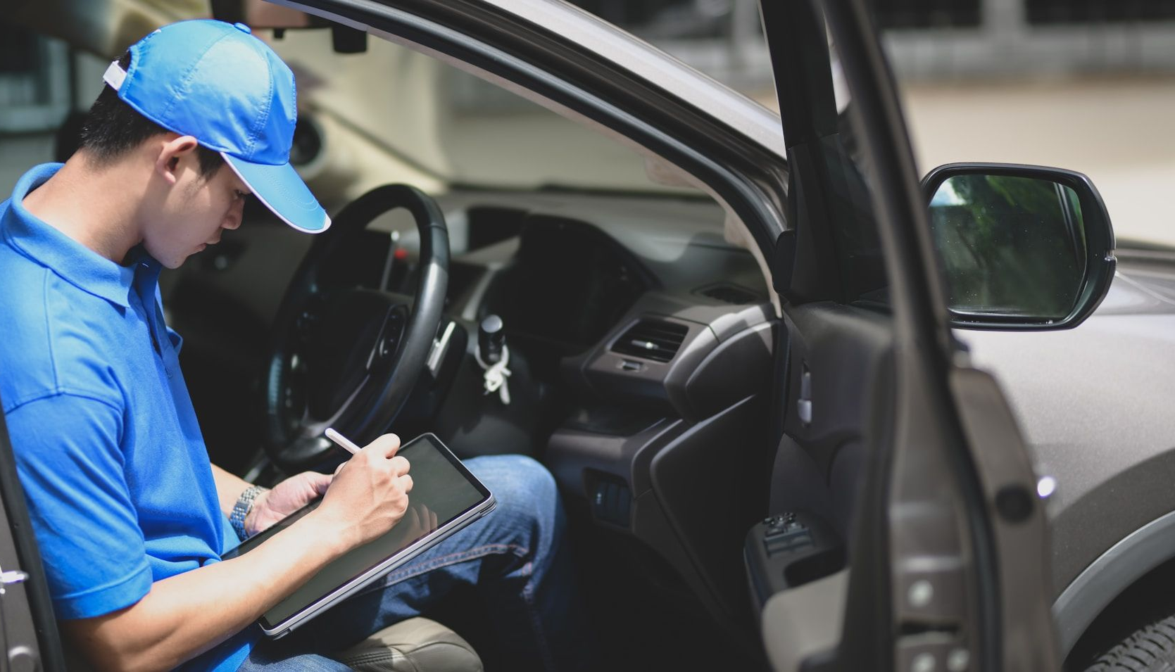 Mechanic in blue uniform writing on a clipboard beside a car with the driver’s door open