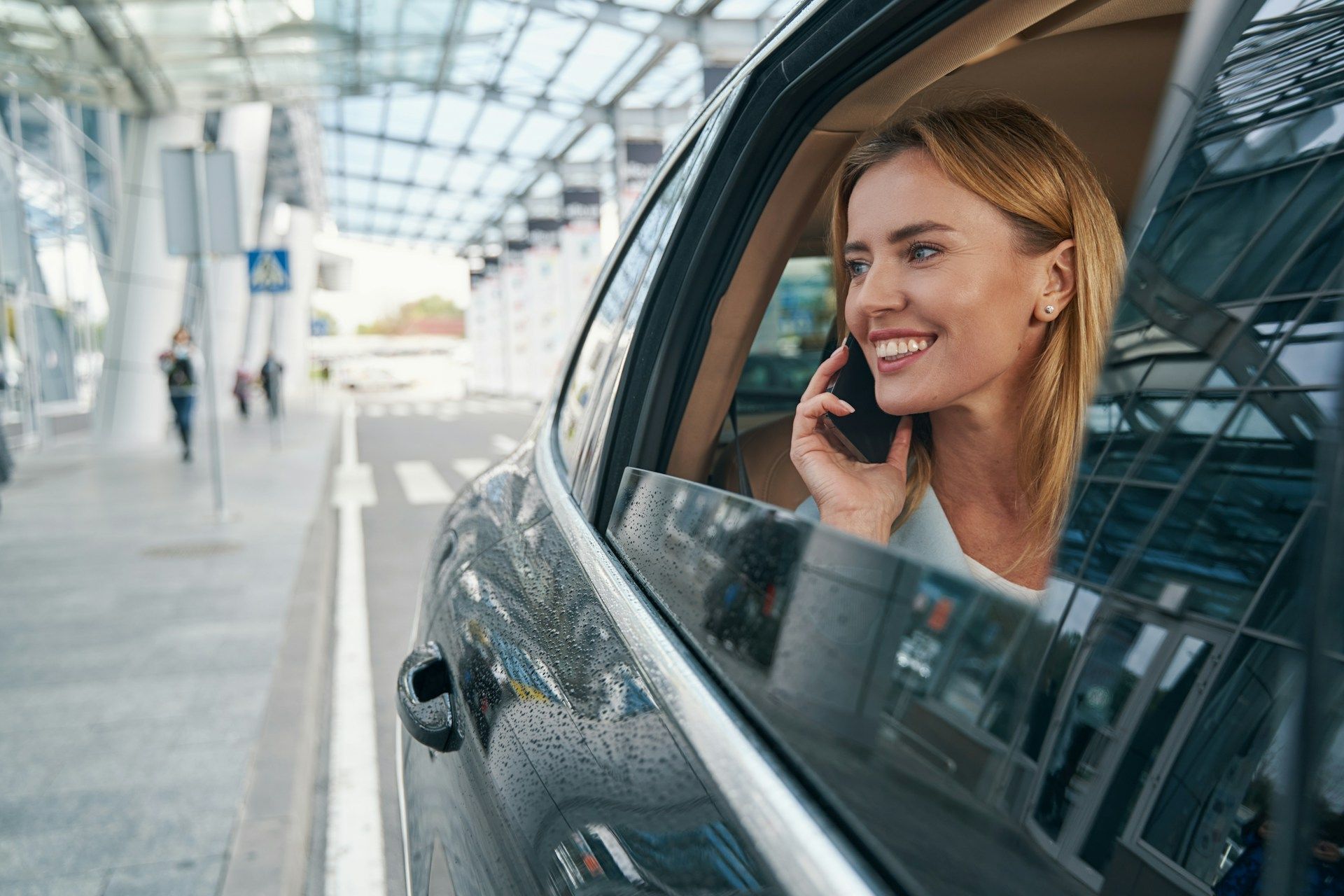 Smiling woman in a car at an airport terminal, talking on a phone through the open window