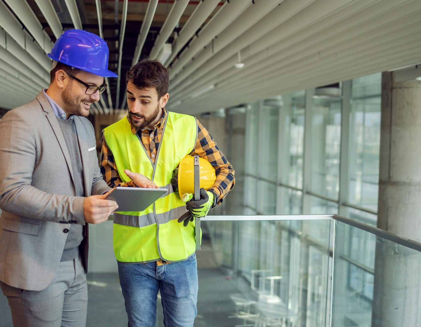 Two construction workers reviewing plans in a modern building interior, one wearing a hard hat and safety vest.