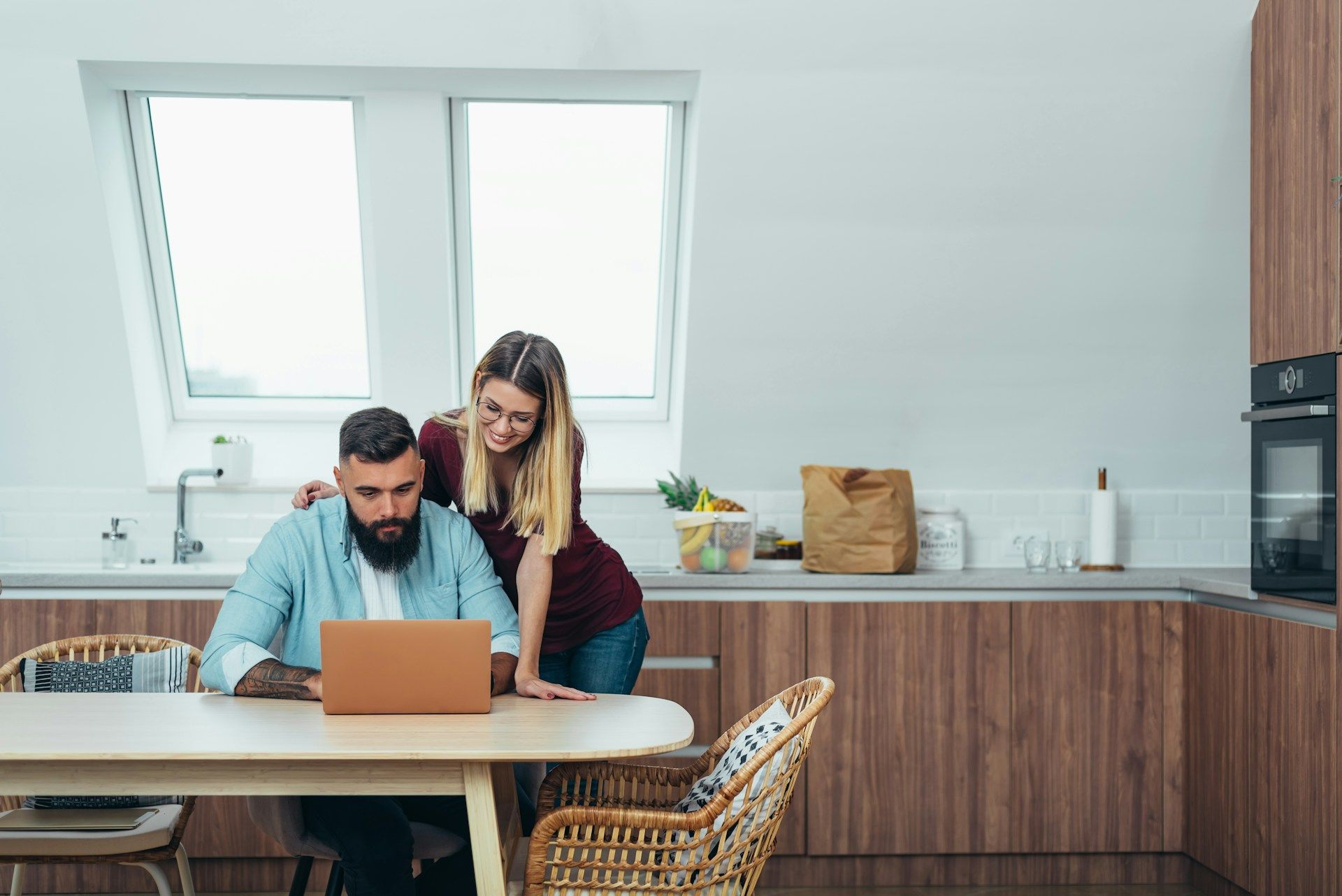 Two people working at a laptop in a bright kitchen with wooden cabinets and a dining table
