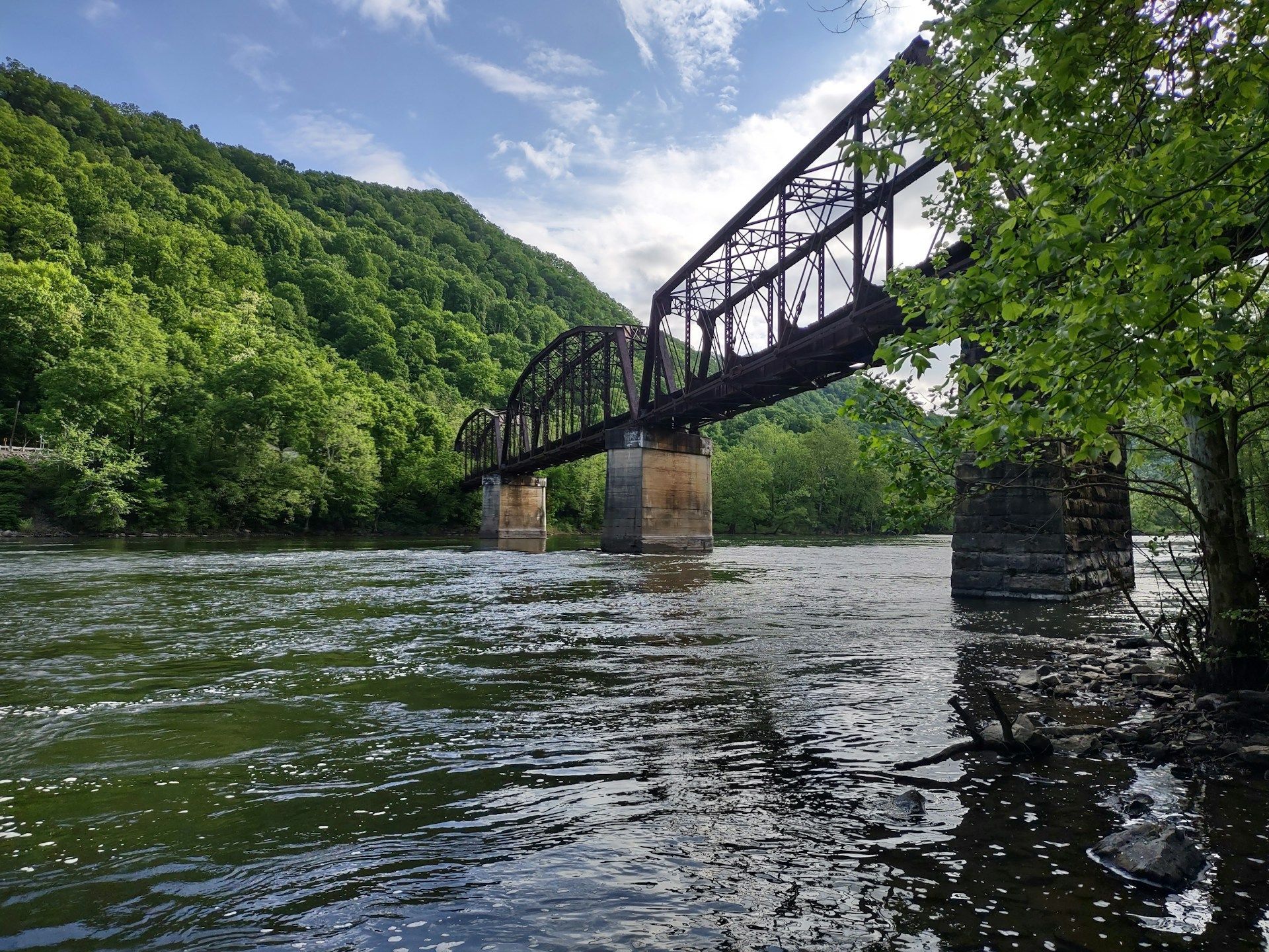 Rusty railway bridge over a river beside lush green forest and a cloudy sky