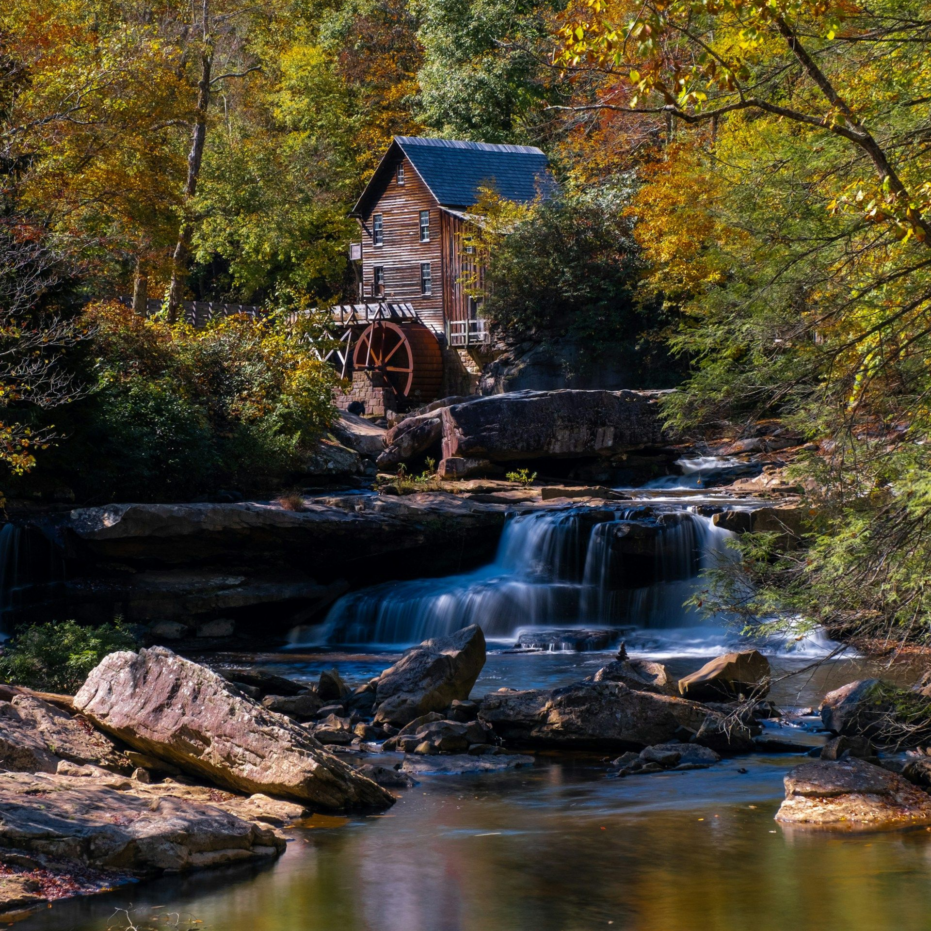 Rustic watermill beside a small waterfall in an autumn forest, with a stream and rocks in the foreground