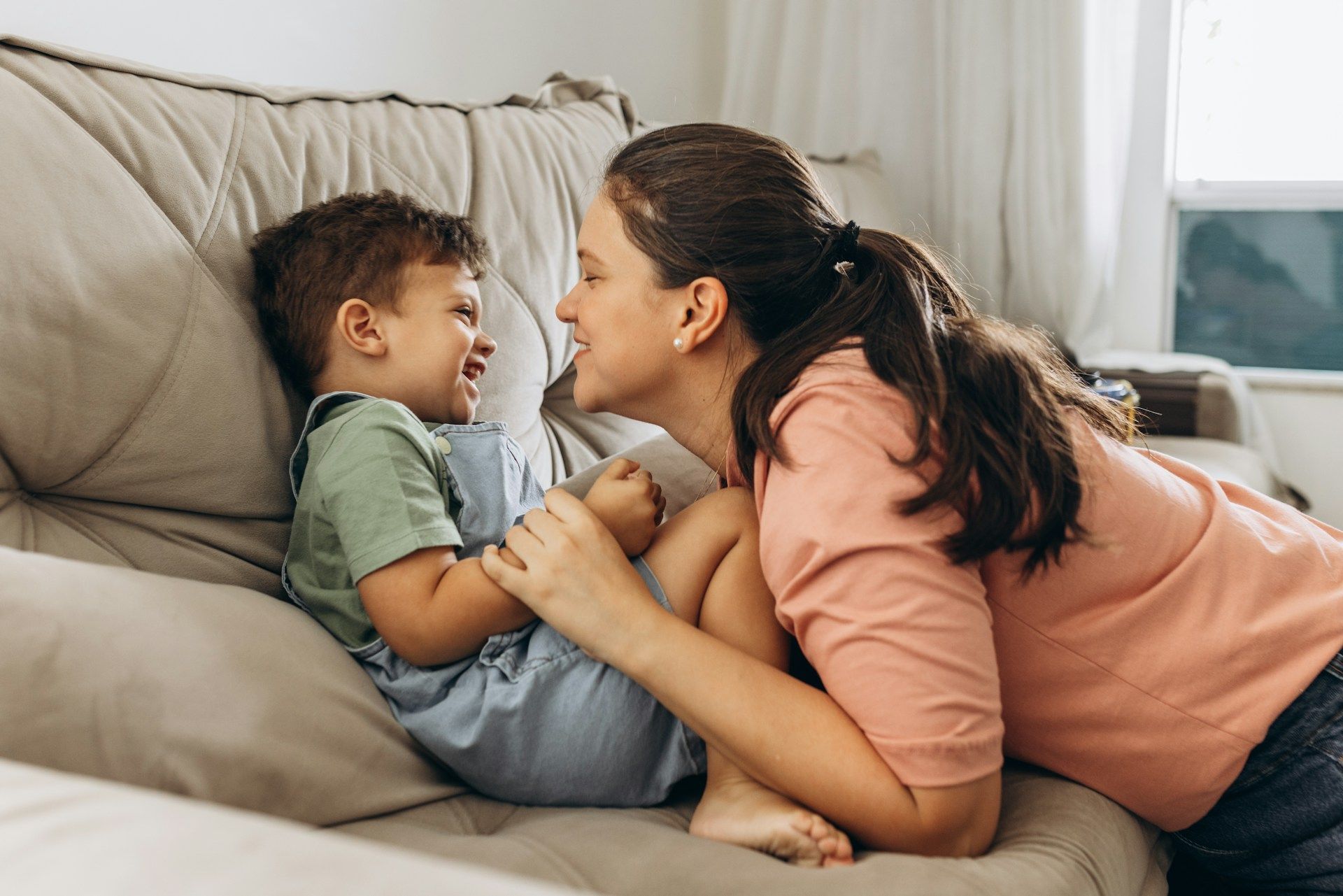Woman lying on a couch smiling at a child, both laughing in a cozy living room