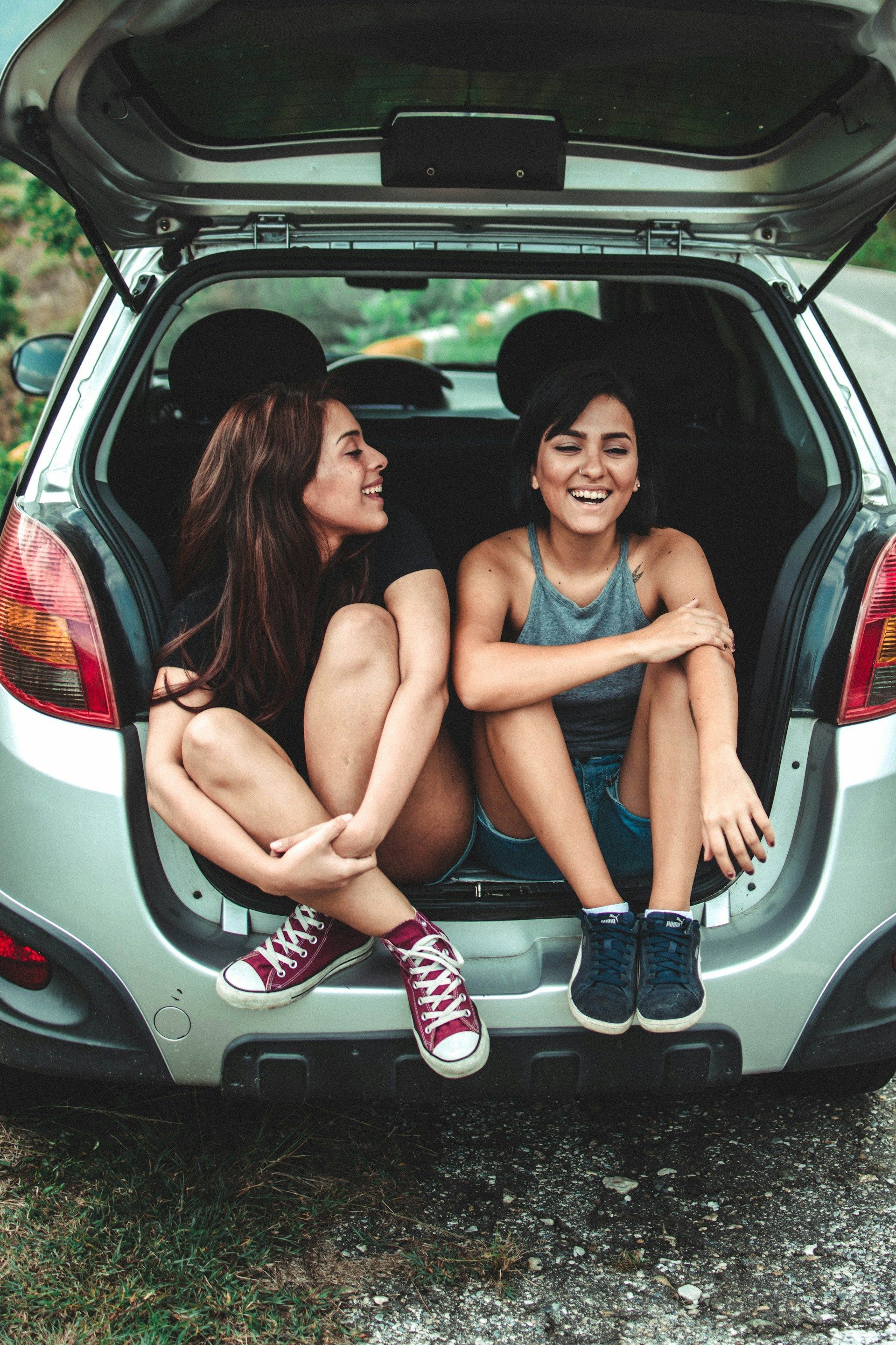 Two people sit in the open trunk of a green car, smiling and chatting outdoors.
