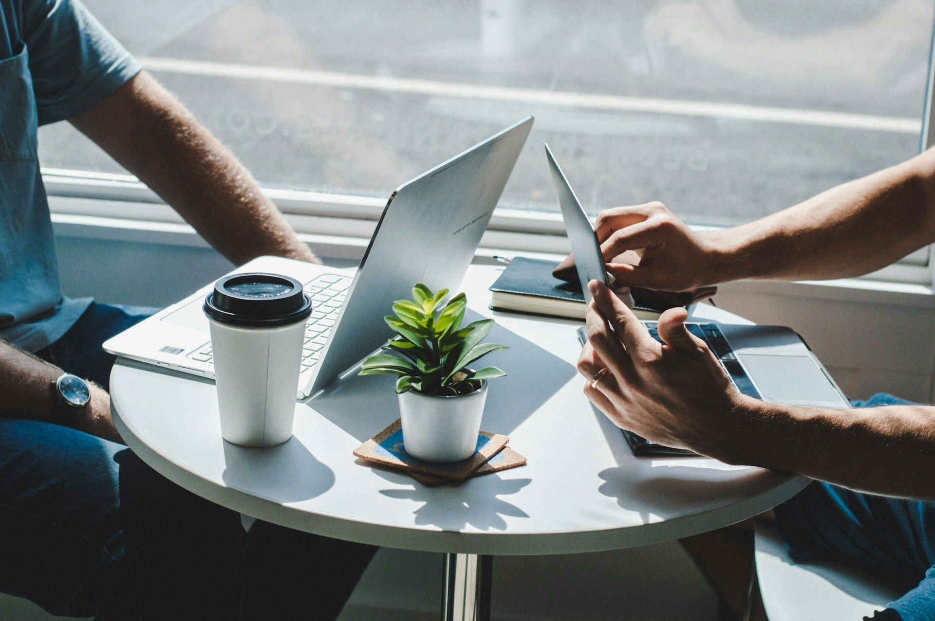 Two people using a laptop at a round table with coffee and a small plant by a window