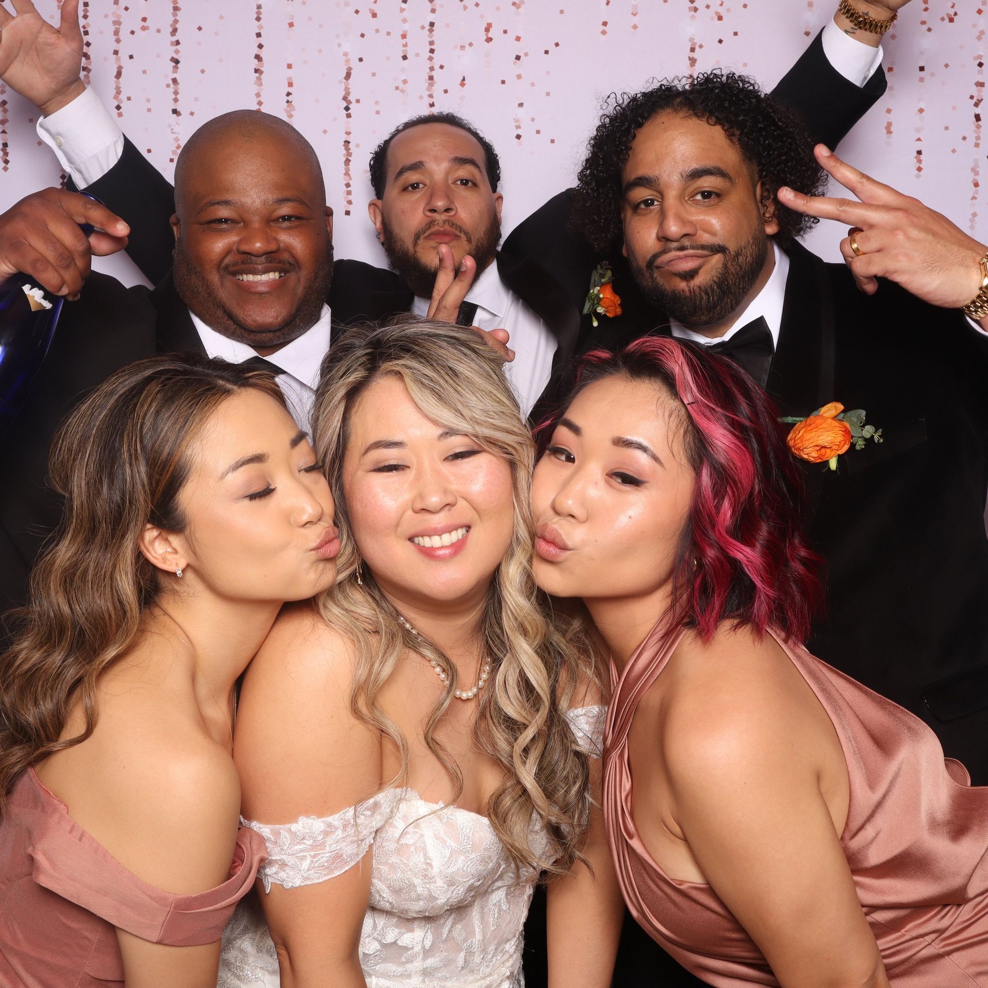 Group posing at a formal celebration, with two women kissing a smiling bride in front and men cheering behind.