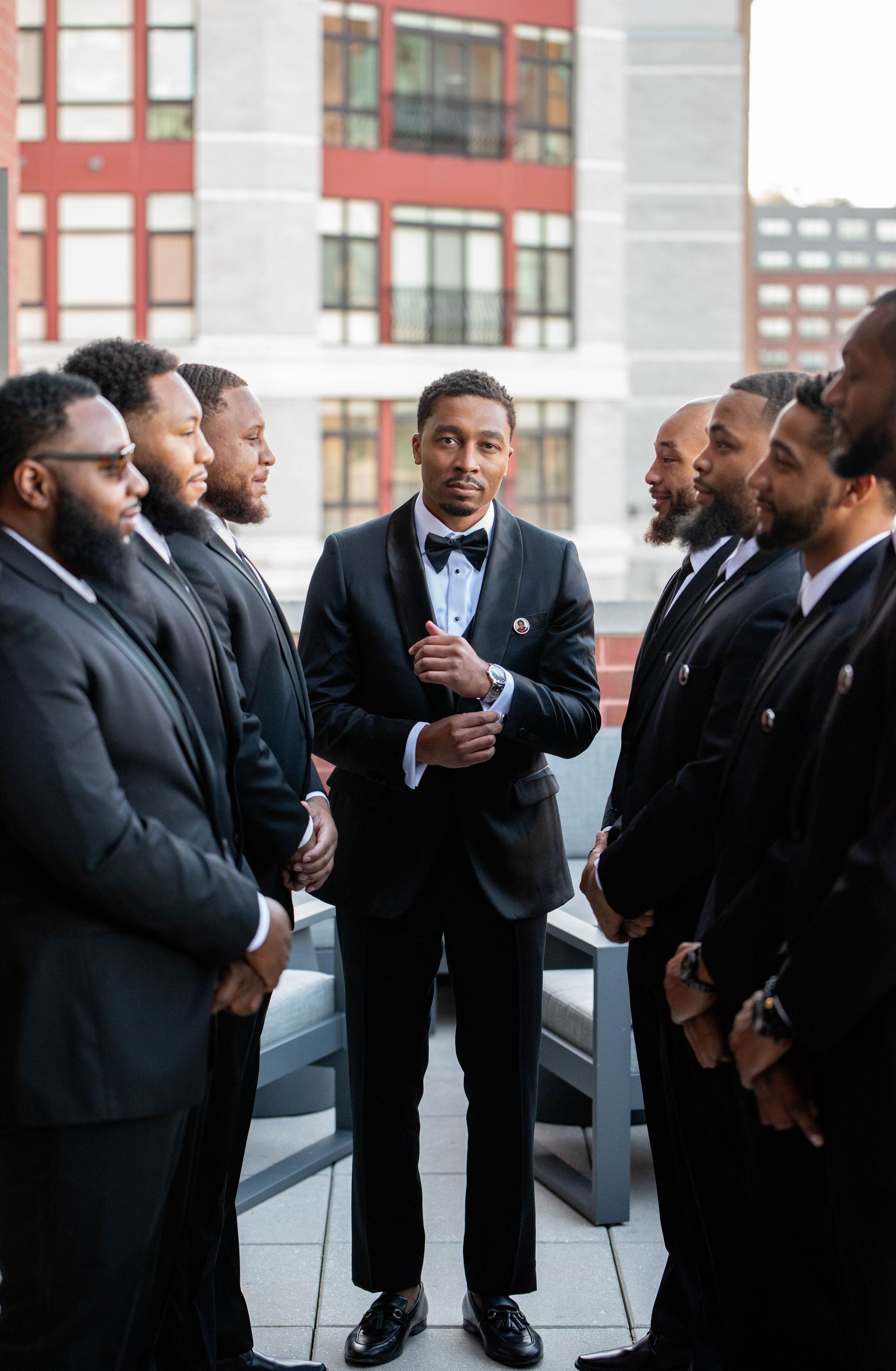 Group of men in black suits standing in a circle outdoors, with one man speaking at the center.