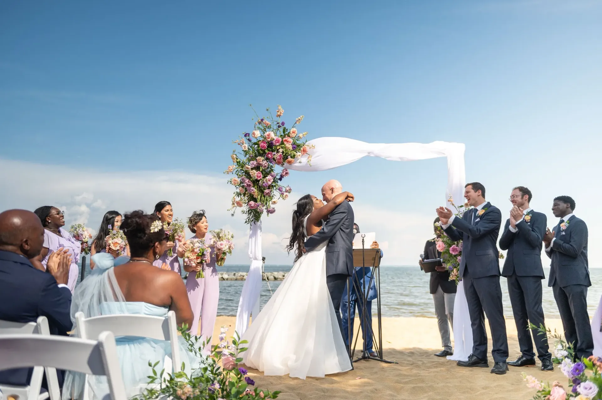 Wedding couple embracing under floral arch on a sunny beach as guests applaud