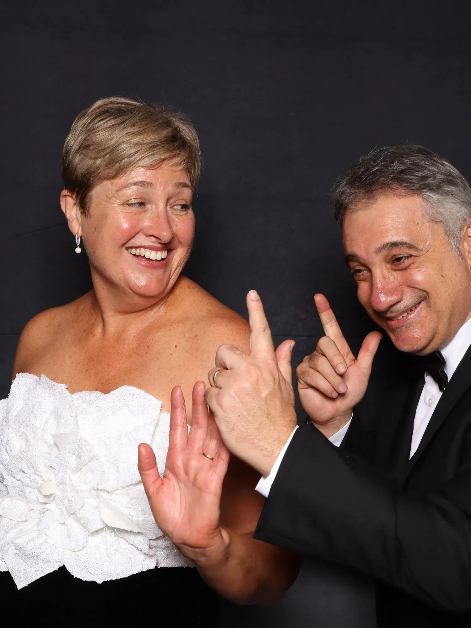 Two people in formal wear smiling and making playful hand gestures at an event