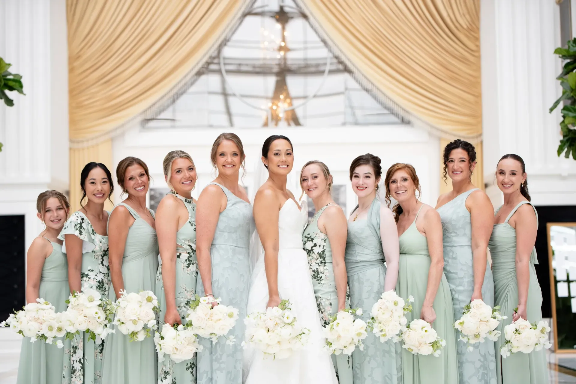 Bridal party in mint dresses posing with white bouquets under gold draped wedding arch