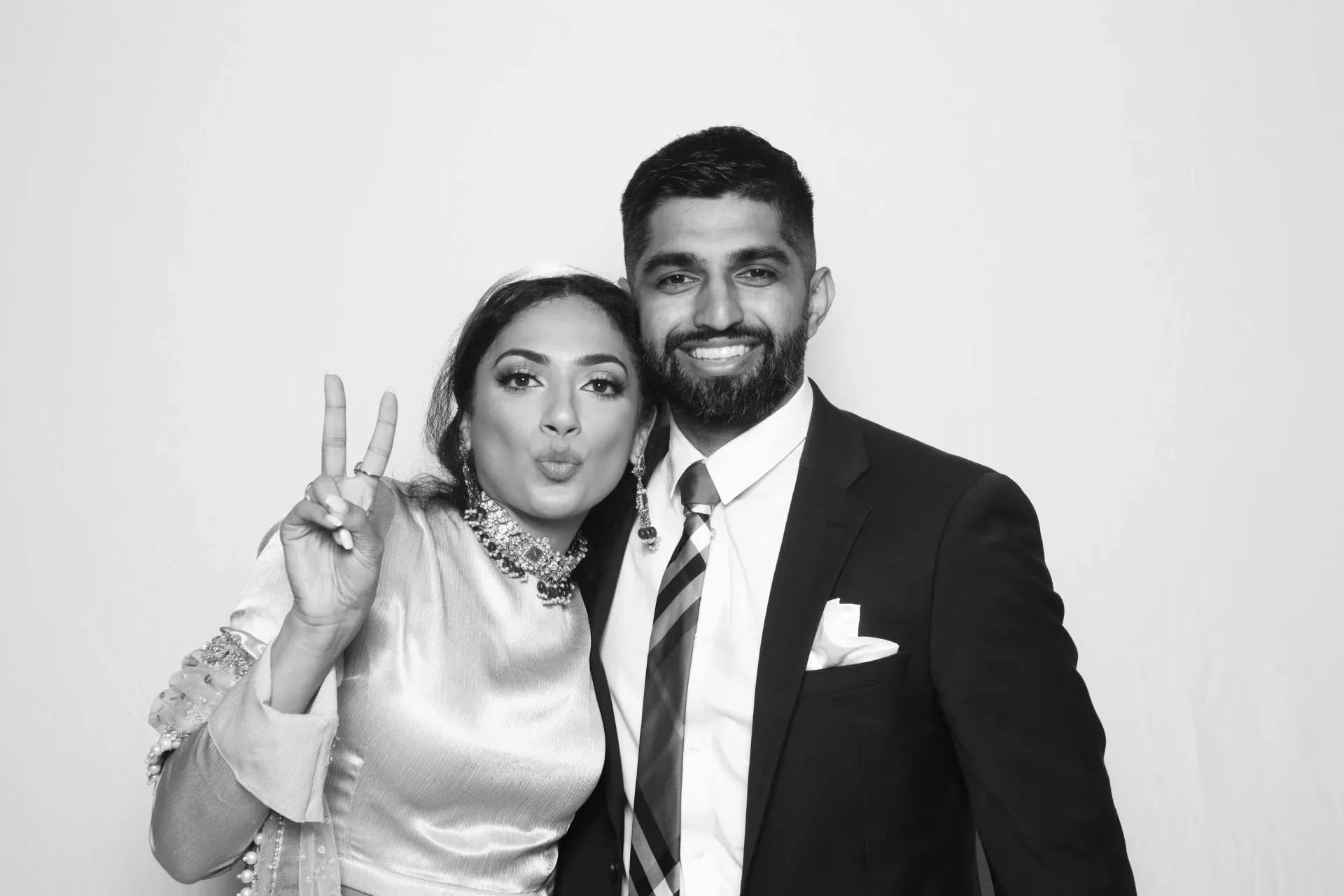 Two people posing in formal attire, smiling and making a peace sign against a plain backdrop in a glam photo booth