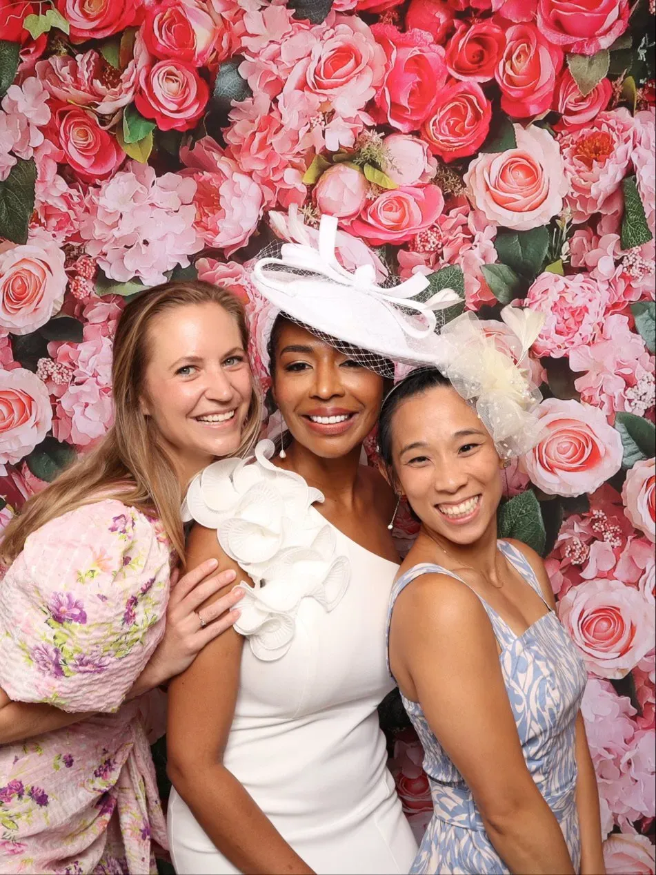 Three women posing in formal dresses before a pink floral backdrop