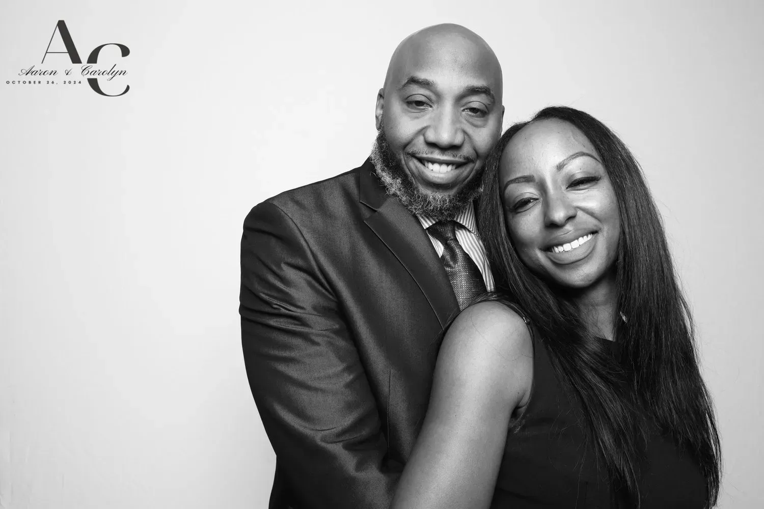 Two smiling people posing closely together against a plain backdrop, one in a suit and one in a sleeveless dress.