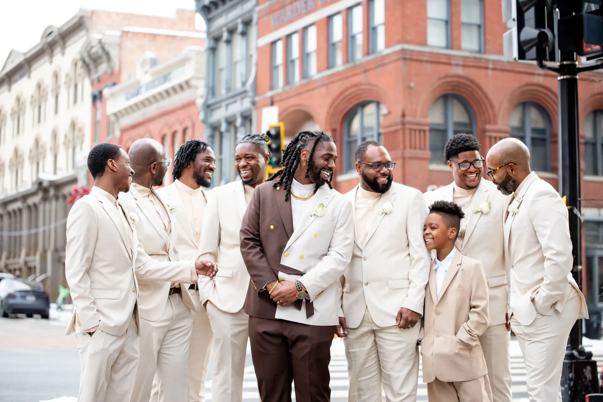 Group of people in cream and brown suits standing together on a city street, smiling and chatting.