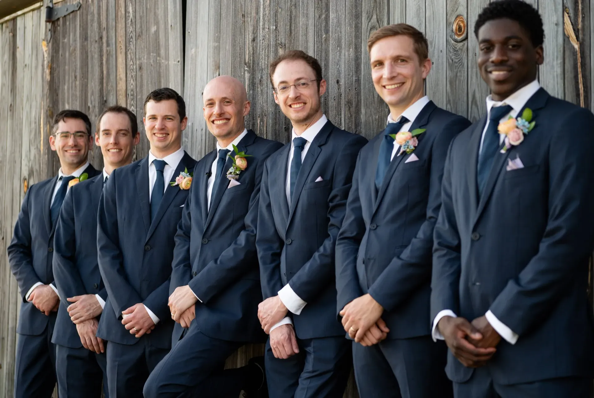 Seven men in navy suits stand in a row outdoors against a stone wall, holding bouquets and smiling.