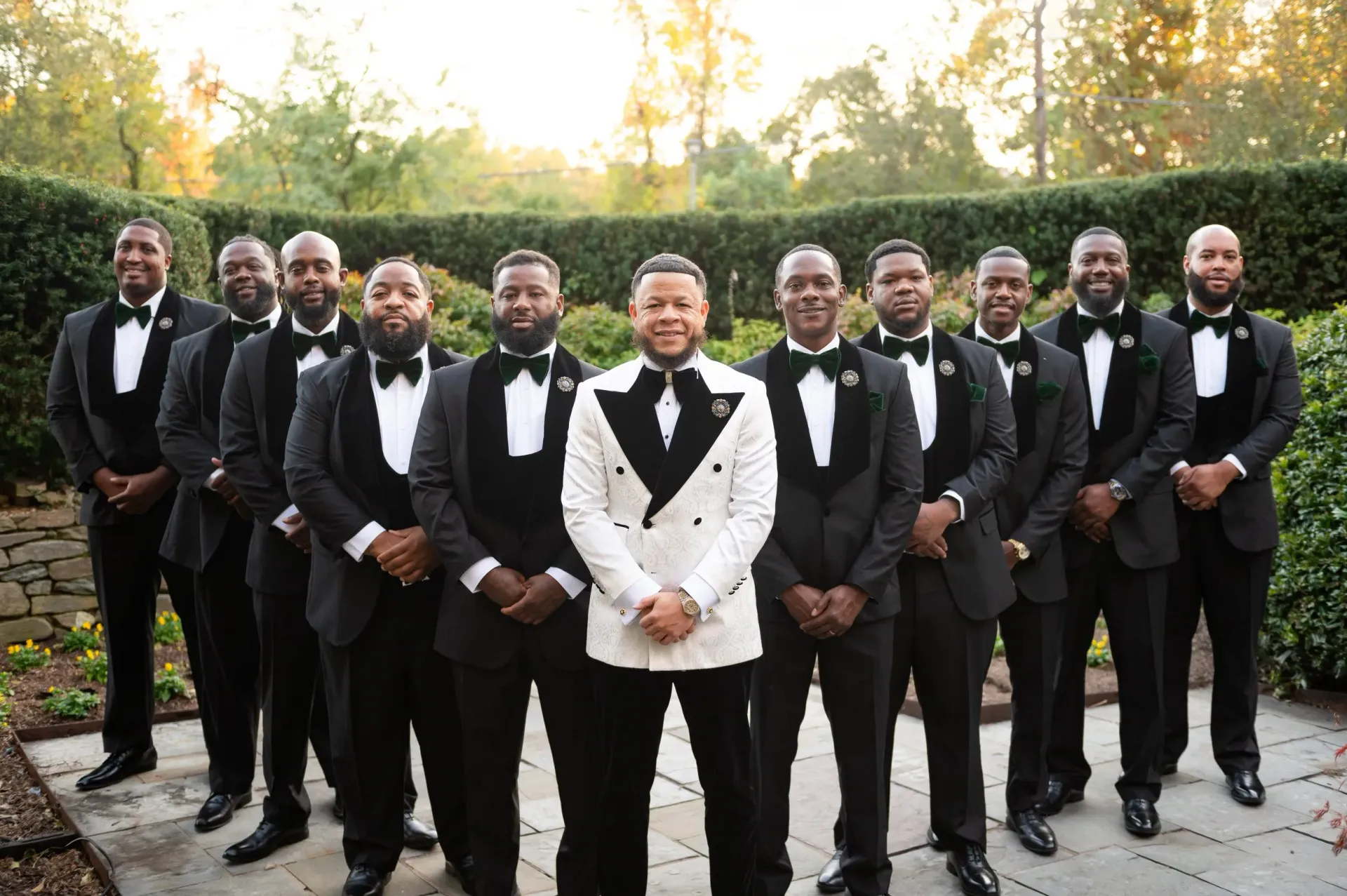 Group of men in tuxedos posing outdoors on a path with autumn trees in the background