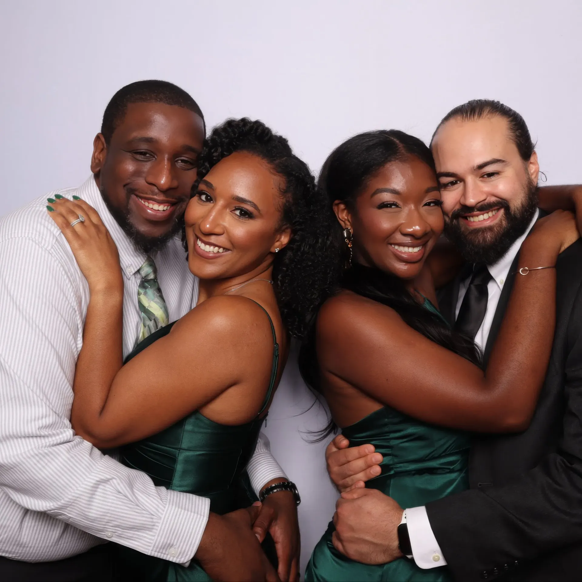 Two smiling couples embracing in formal attire against a light backdrop