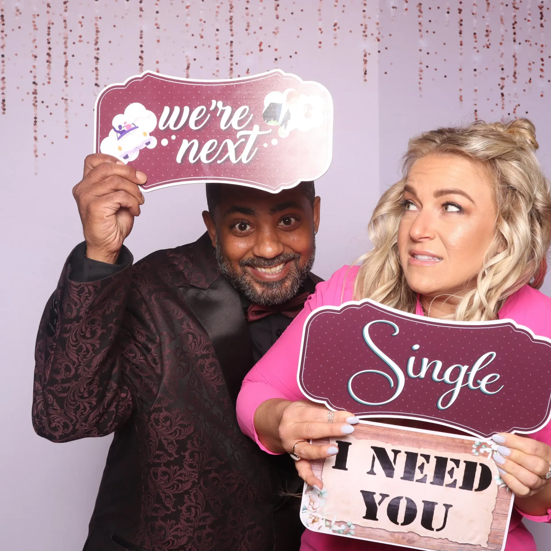 Two people holding wedding-themed signs: “we’re next,” “Single,” and “I NEED YOU” against a pink backdrop