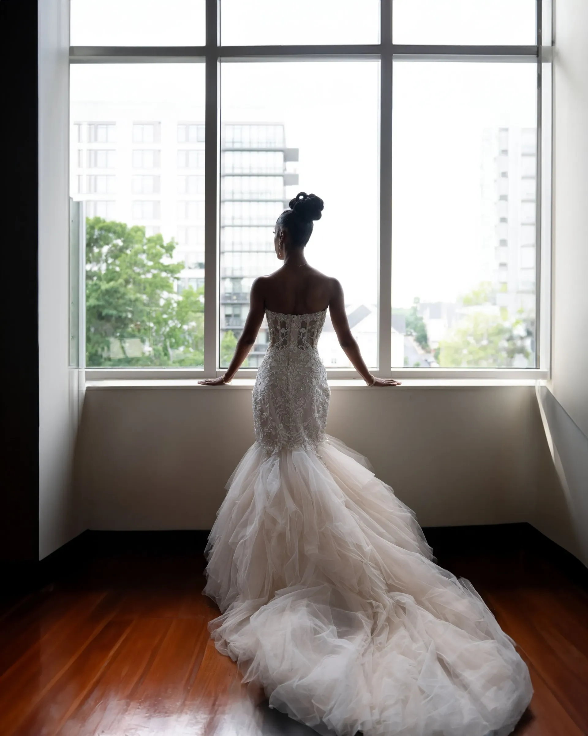 Bride in a white lace gown standing by a bright window, looking out over the city.