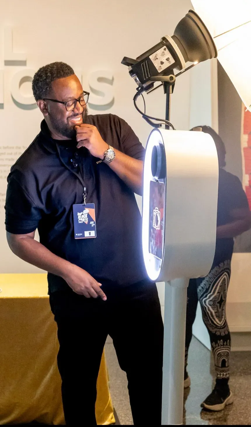 Man speaking into a microphone beside a glowing photo booth in a hallway