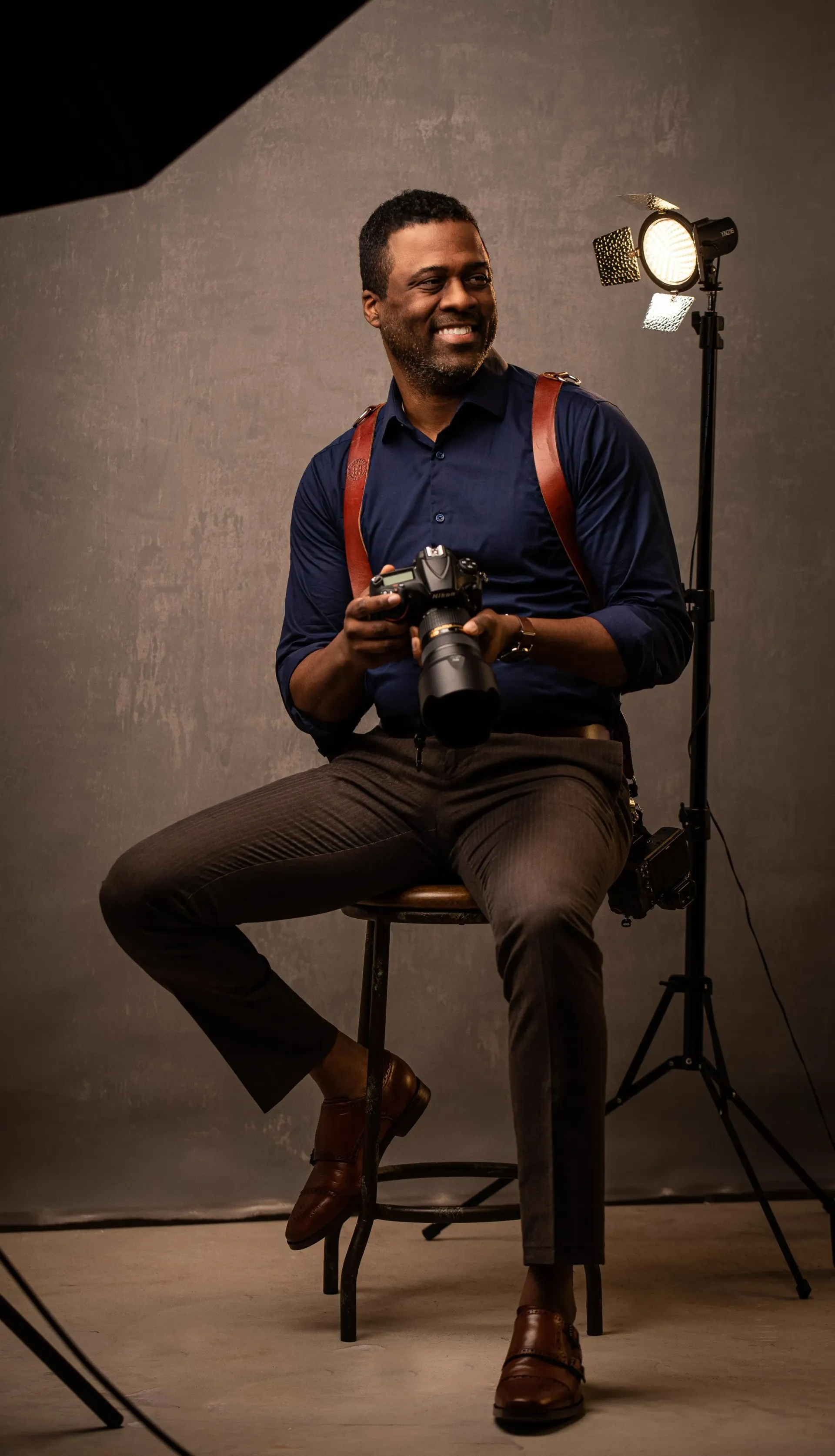 Man seated on a stool in a studio, holding a camera and smiling beside a light stand.