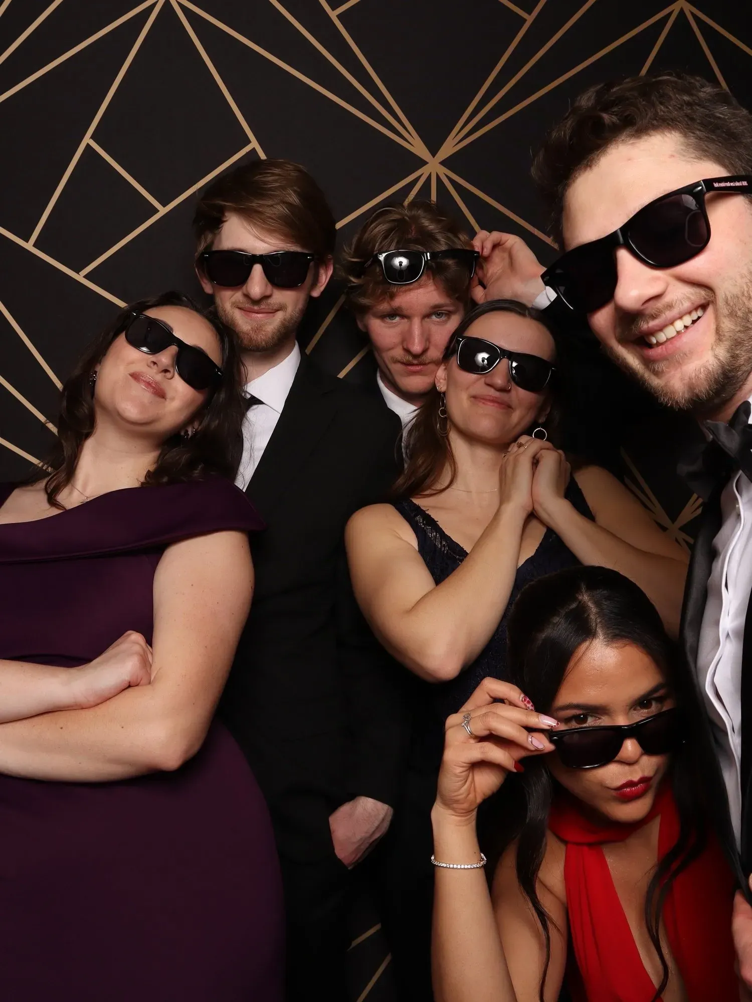 Five people wearing sunglasses pose closely together against a black-and-gold geometric backdrop.