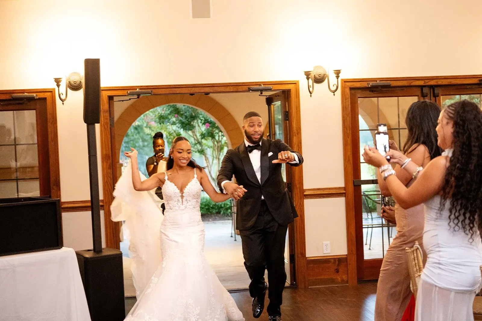 Bride and groom entering a reception hall, smiling and waving as a guest takes a photo.