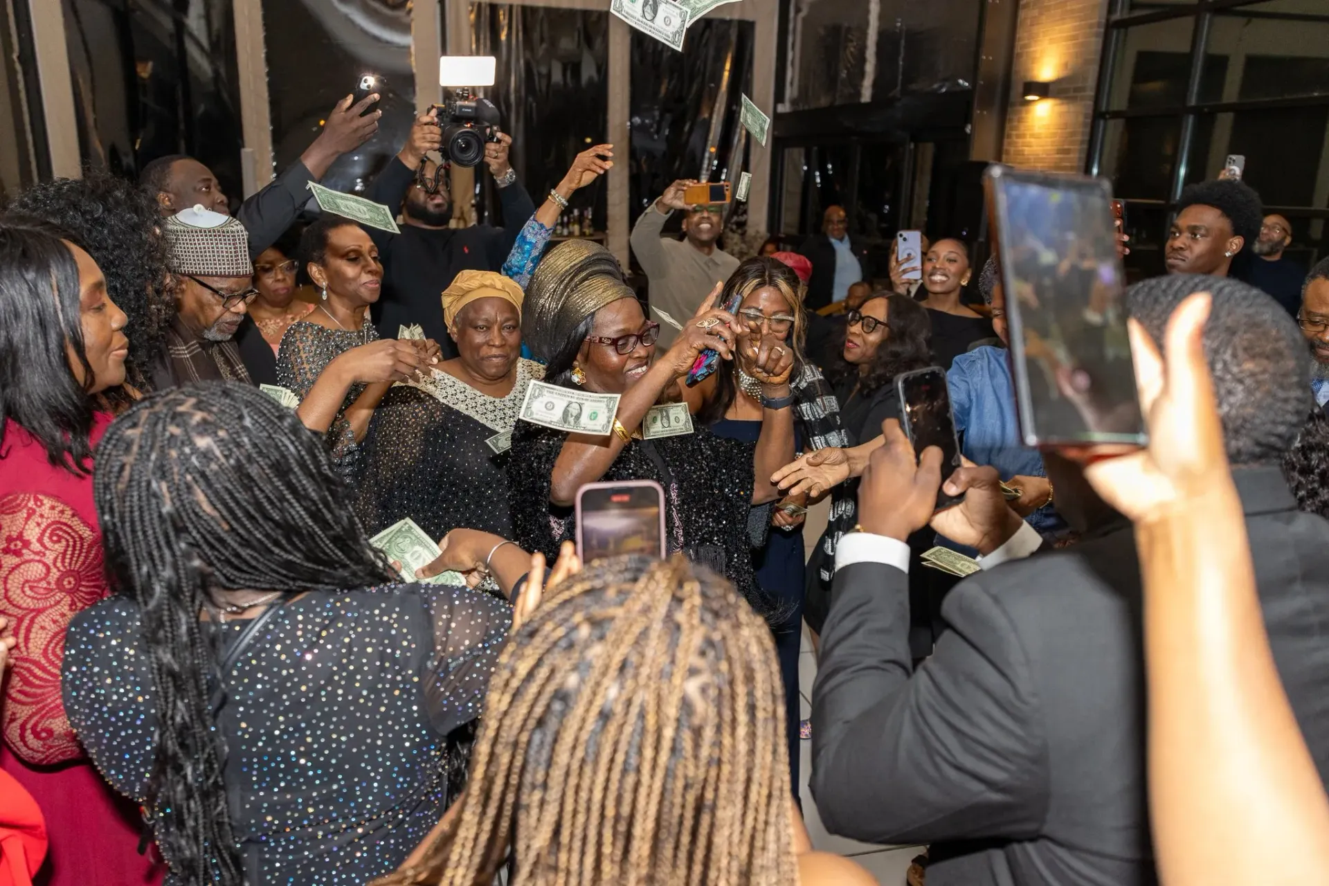 Crowd at an indoor party cheering and raising phones for photos, with lights and decorations in the background