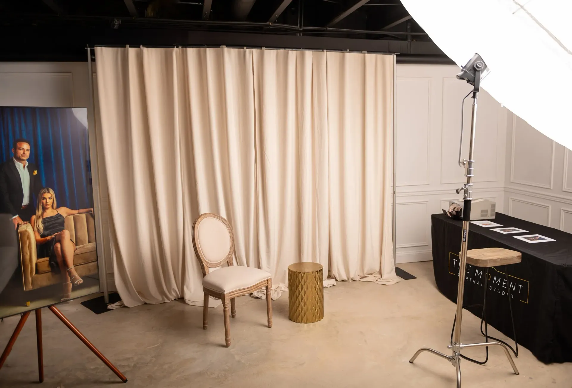 Studio set with cream curtains, white chair, small table, and lighting equipment on the right.