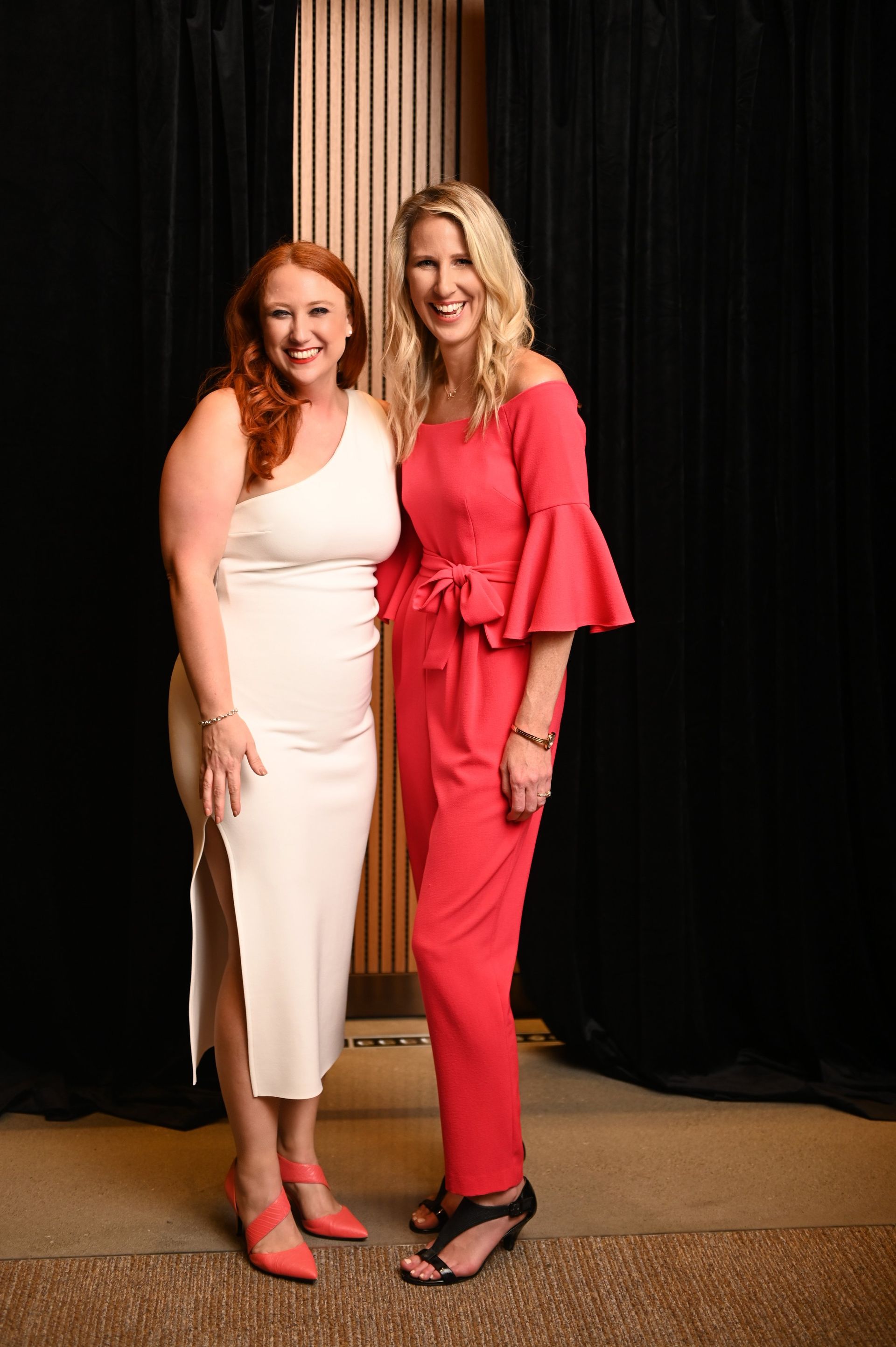 Two women posing together in front of a black curtain, wearing white and pink outfits.