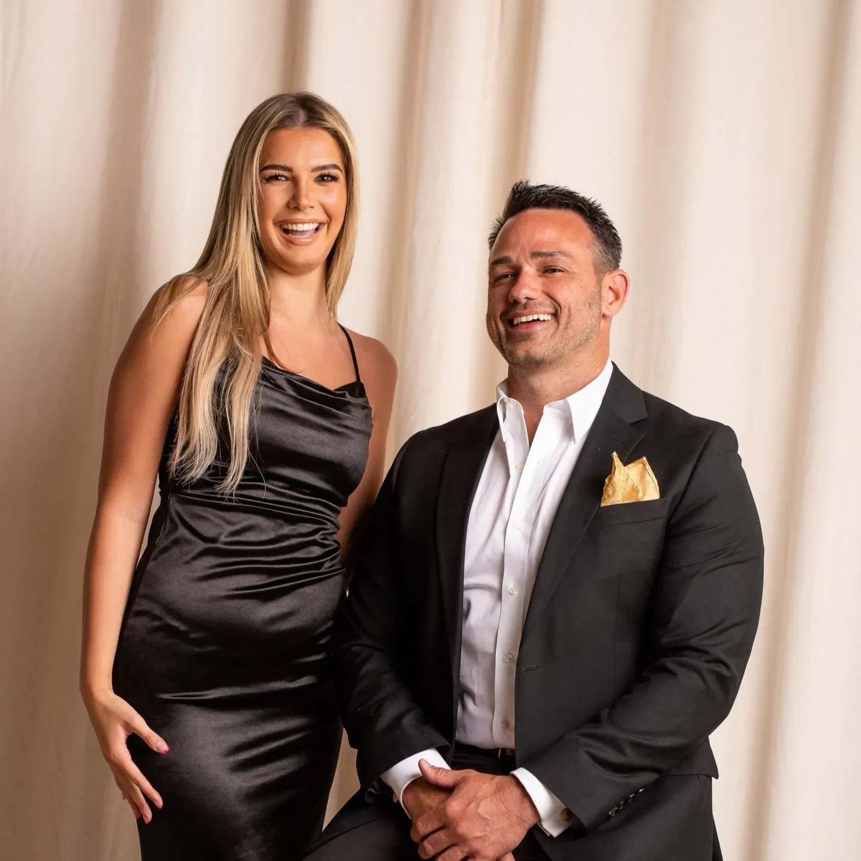 Two smiling people posed in formal black and white attire before a beige curtain backdrop.