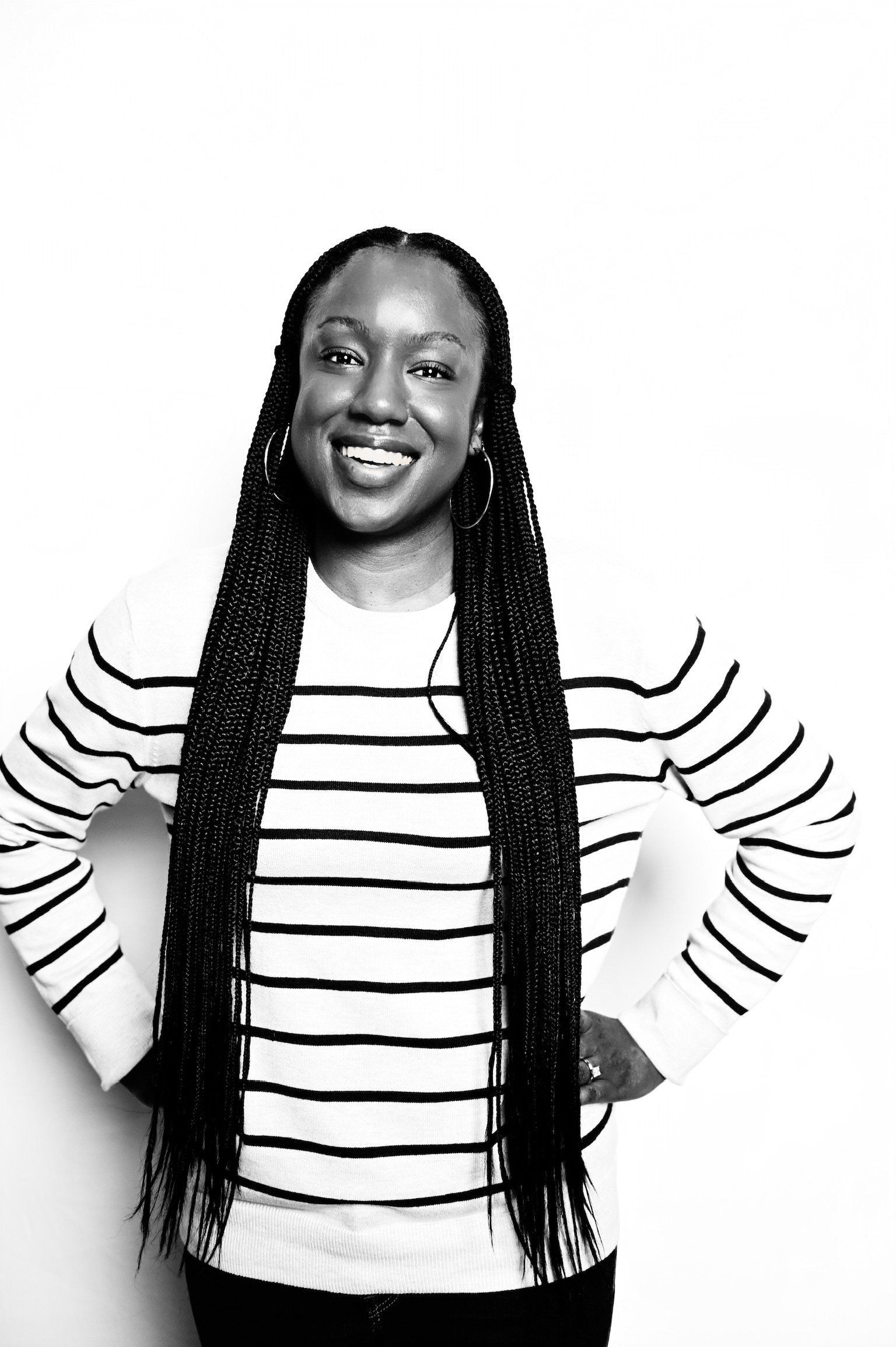 Smiling person in a black-and-white striped shirt with long braids, standing with hands on hips.